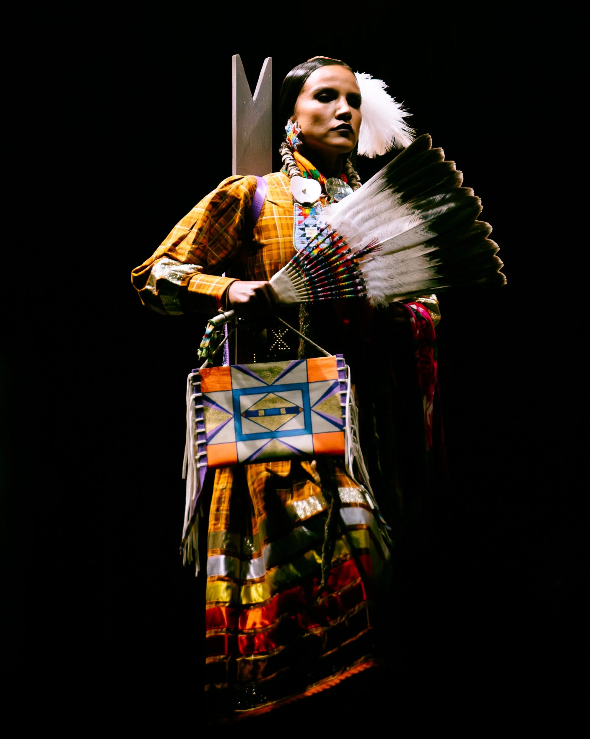 Articulated mannequin dressed in Native American inspired clothing and jewelry, holding a feather fan and a colorful woven bag, against a black background.