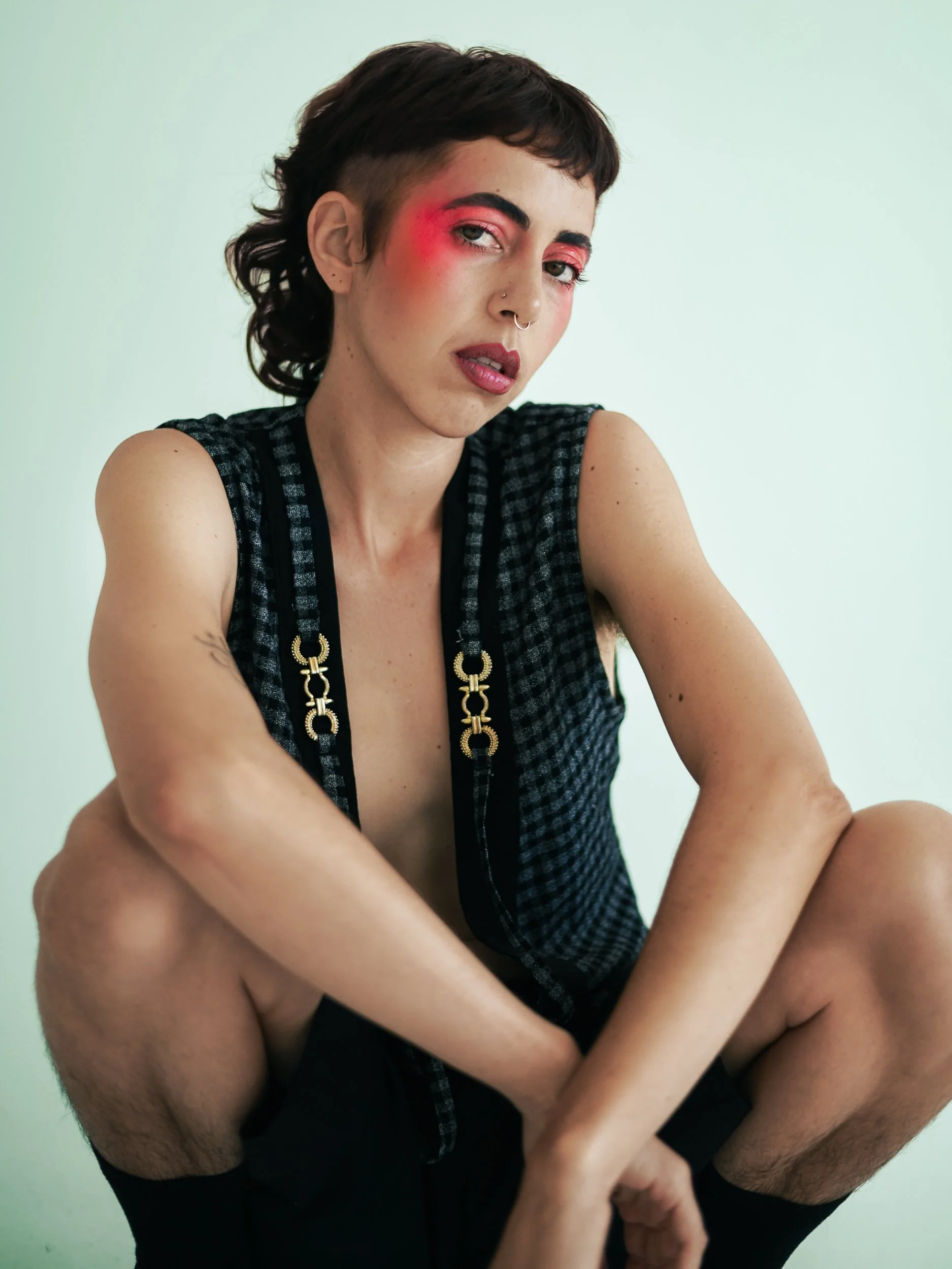 A person with short dark hair, bold makeup including pink eyeshadow and lipstick, a nose ring, and a tattoo on their left arm, wearing a sleeveless checkered top with gold chain details, posing against a plain white background, photographed during a 