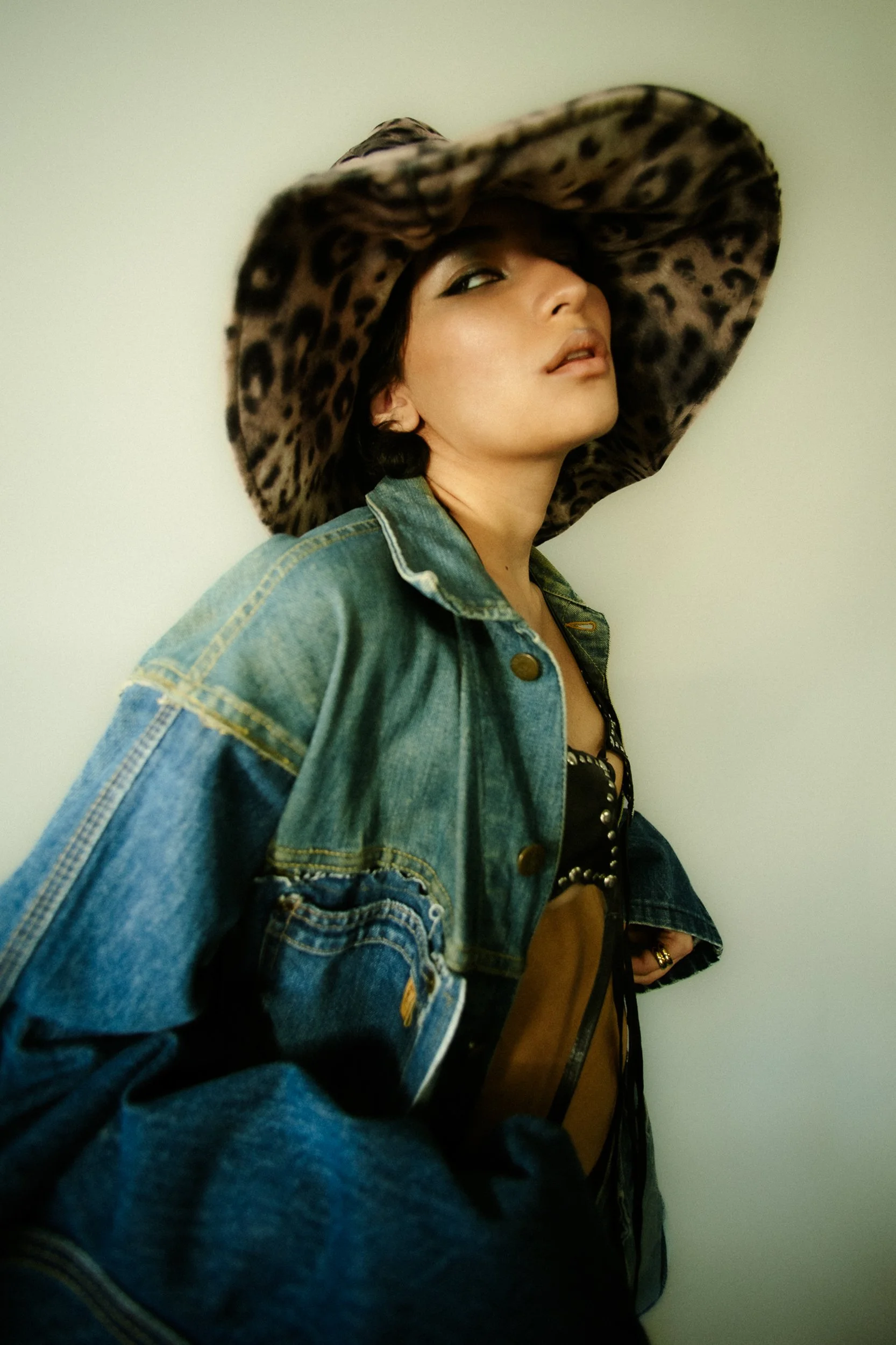A woman wearing a large leopard print hat, denim jacket, and a black top with silver studs, leaning against a white wall with a confident expression, photographed during a fashion editorial photography session in Los Angeles with fashion photographer