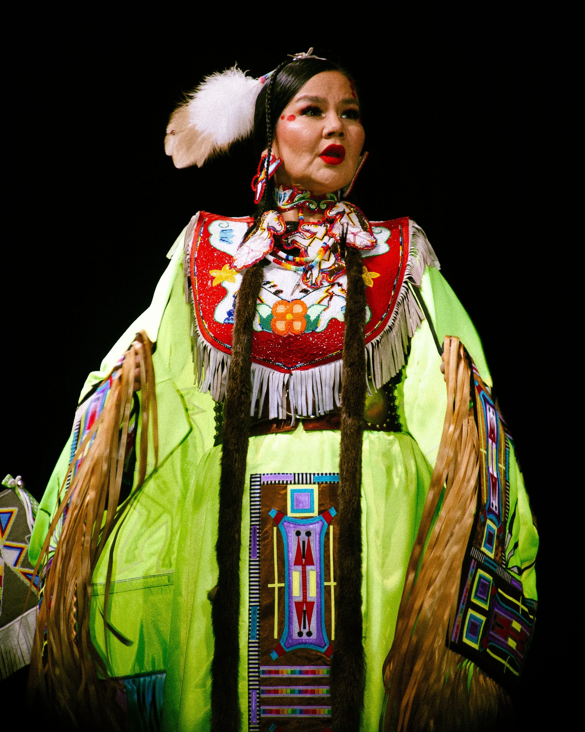 A woman dressed in traditional Indigenous ceremonial attire, with intricate beadwork, fringes, and bright colors, standing against a black background.