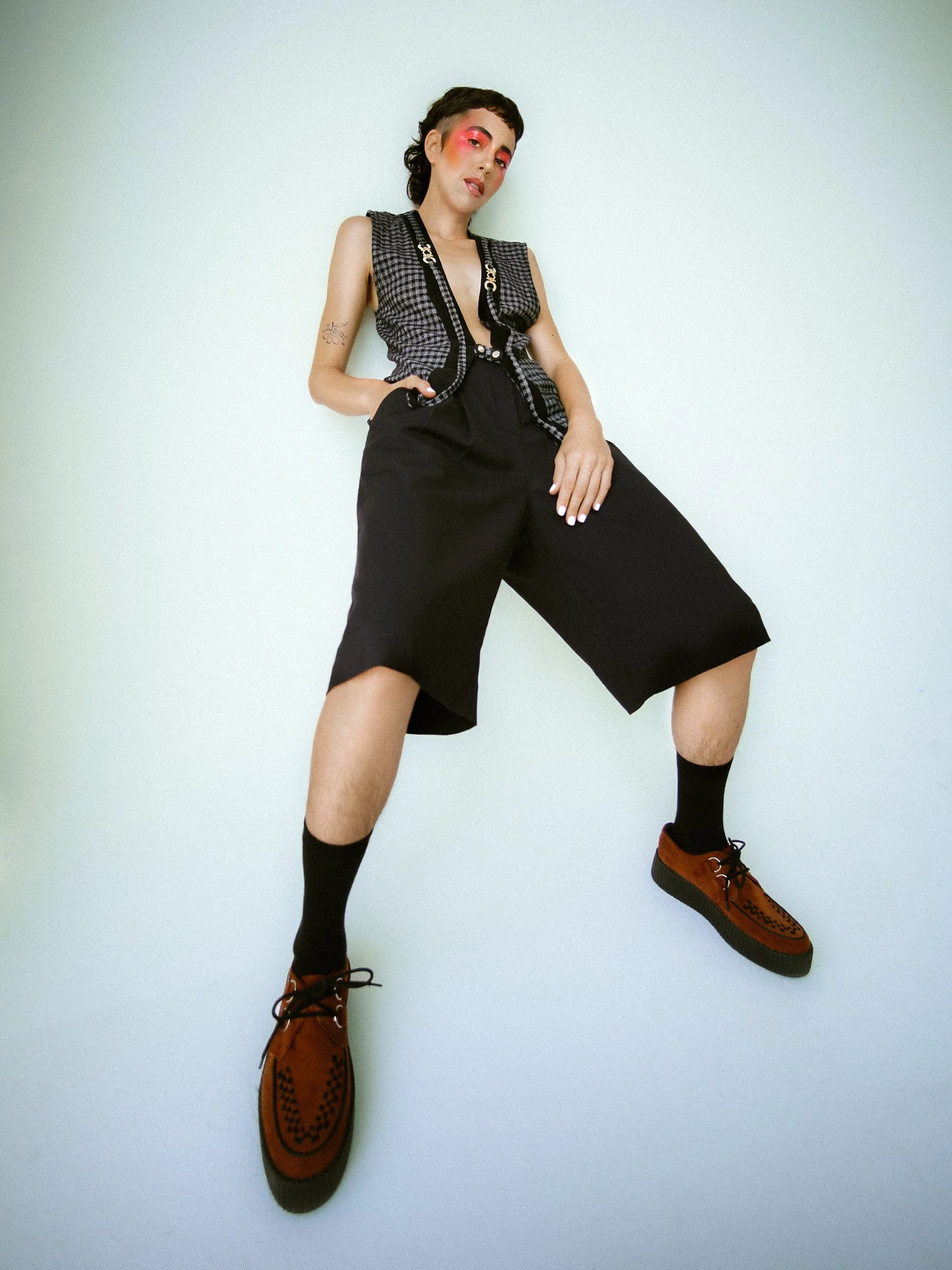 A young person with short black hair, pink eyeshadow, and makeup, wearing a black and white patterned vest, loose black shorts, black socks, and brown shoes, standing against a plain light background, photographed during a studio photography session 
