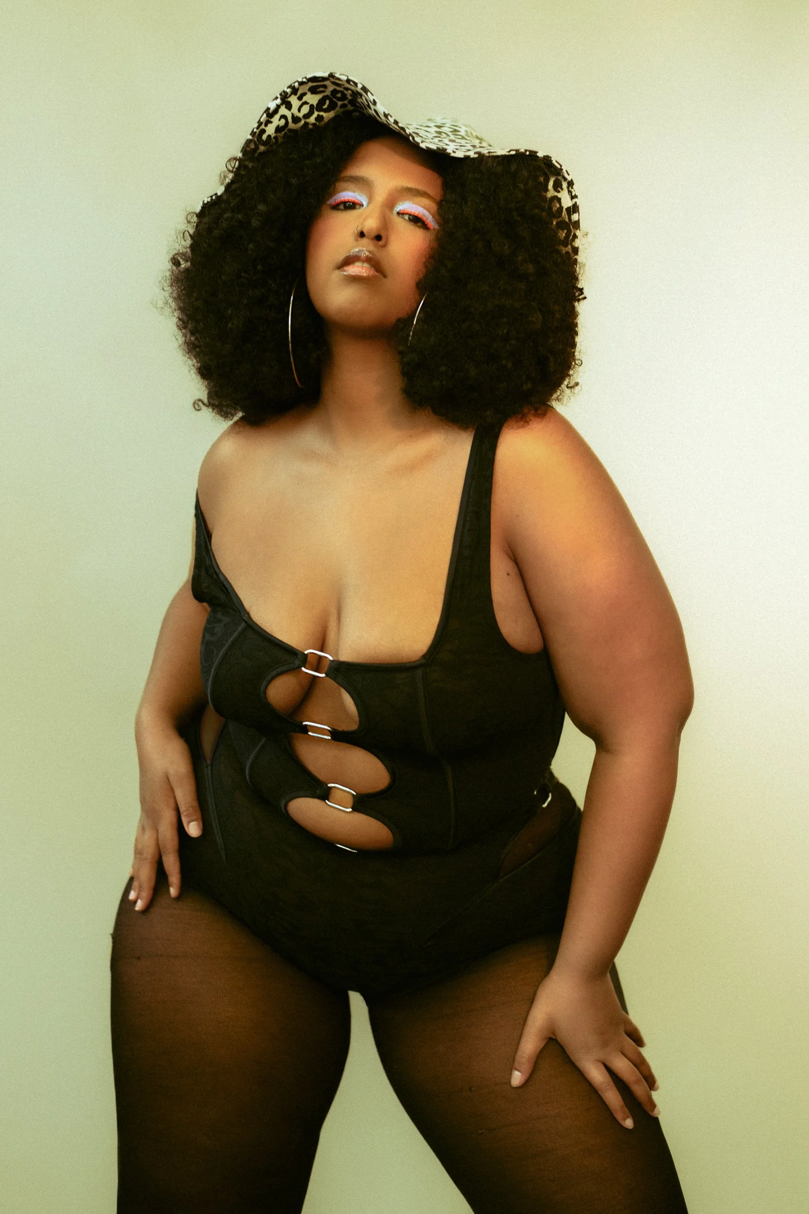 A woman with curly black hair wearing a leopard print hat, large hoop earrings, an eye-catching black top with cut-outs, and sheer black stockings, standing confidently with one hand on her hip against a plain background, A woman with curly black hai