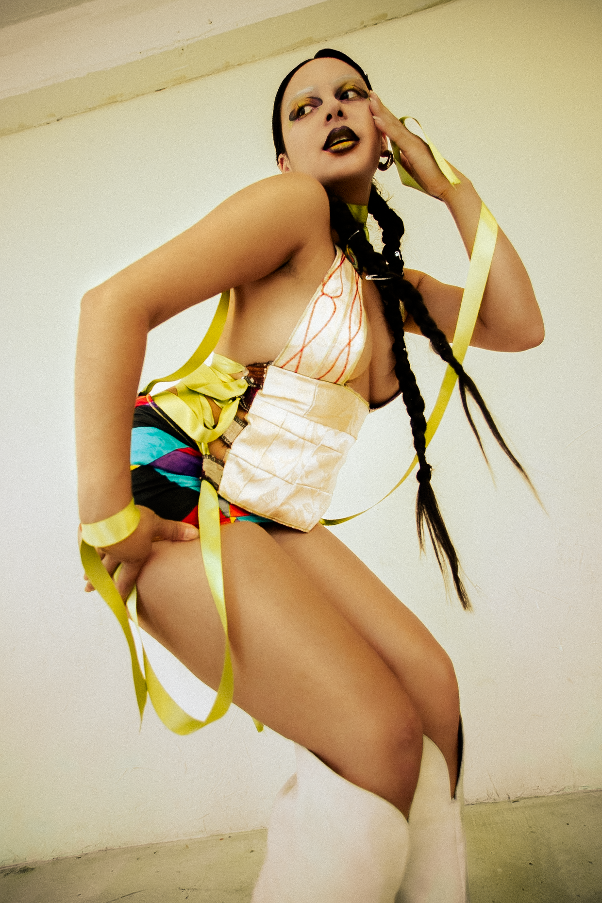 Person with braided hair wearing colorful, bold makeup and a creative, fashion-forward outfit, posing indoors against a plain white wall and glossy concrete floors, shot for an editorial fashion photography session in Los Angeles with fashion photogr