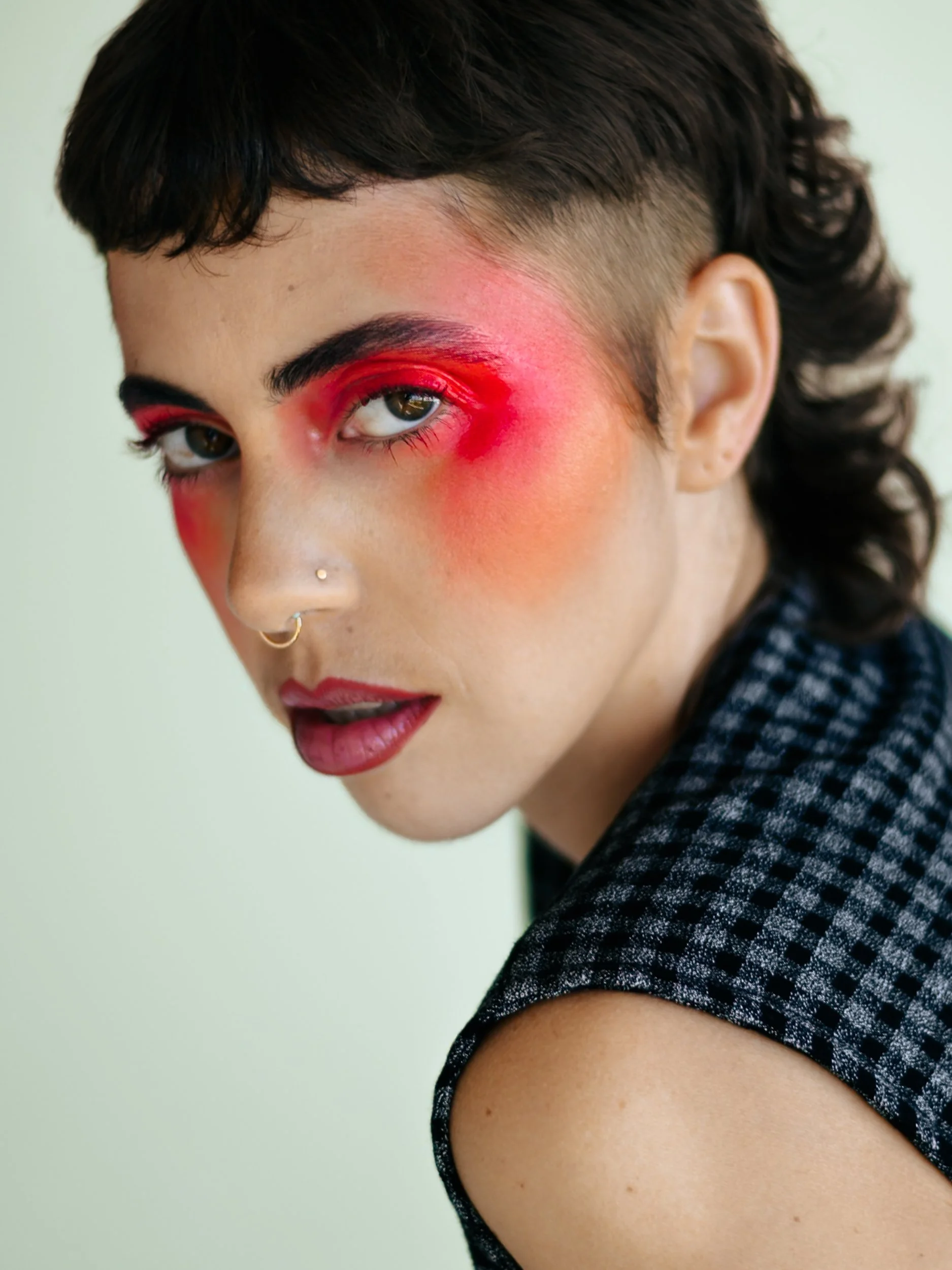 A person with short dark hair, green eyes, and multiple facial piercings wearing vibrant makeup with red and pink eyeshadow and lipstick, dressed in a black and white checkered top, photographed during a studio photography session in Los Angeles for 