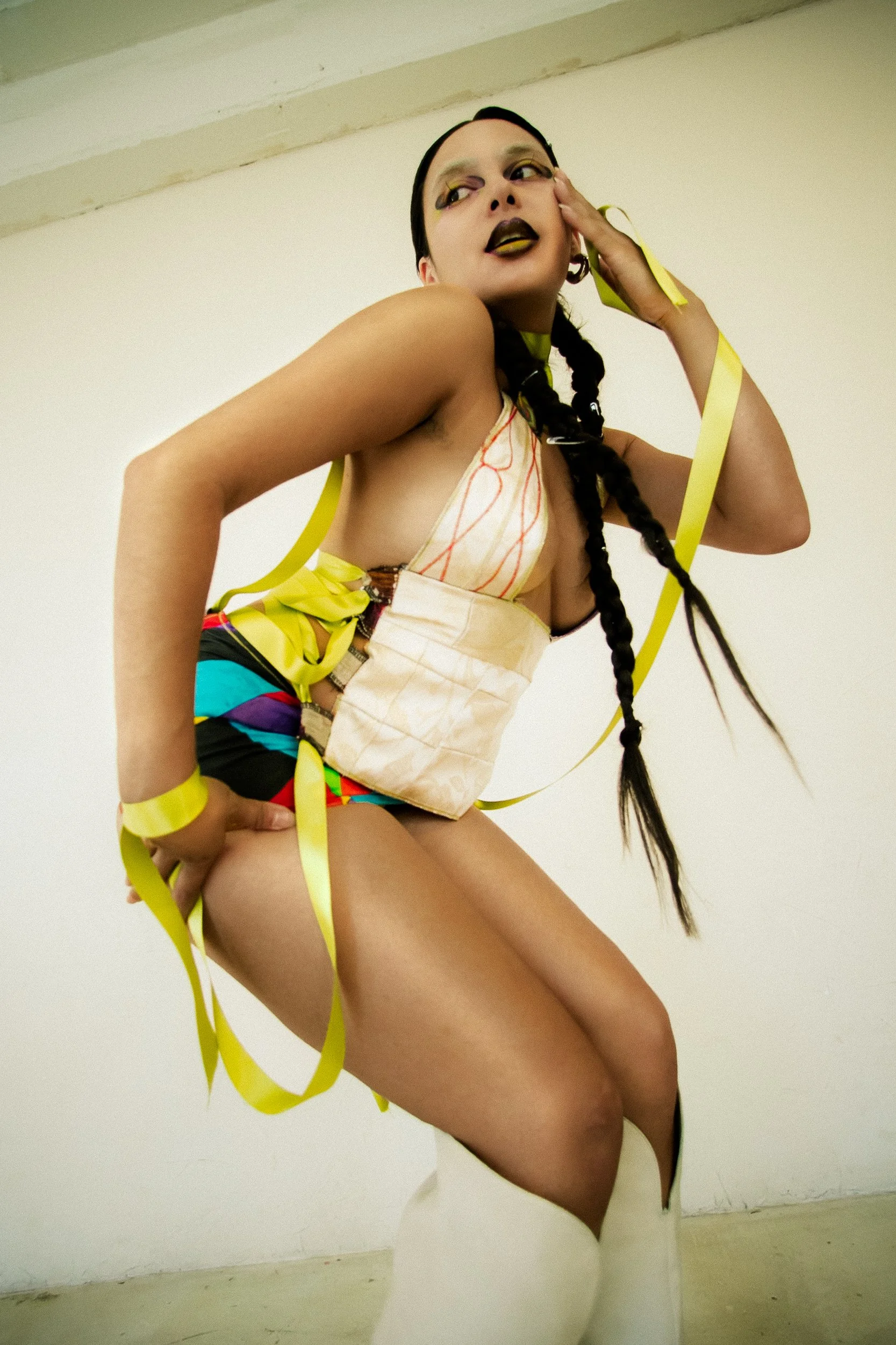 A tan woman model with long black braided hair, wearing colorful shorts and white boots, with yellow ribbons and a padded beige top, striking a pose against a plain wall, photographed during a fashion editorial photography session in Los Angeles with