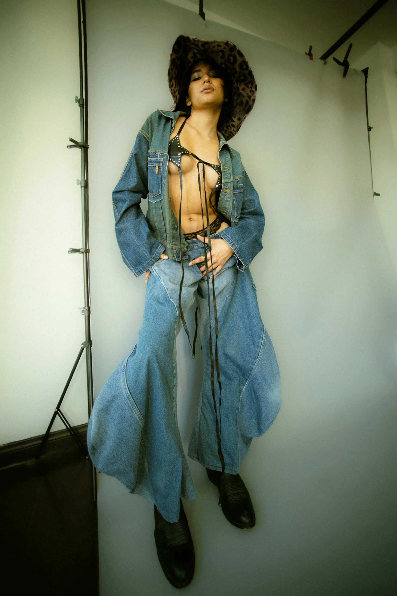 Model wearing a wide-brim hat, open denim jacket, black lingerie, and black shoes, posing against a plain background, photographed during a fashion editorial photography session in Los Angeles with fashion photographer Gí Tenamá..