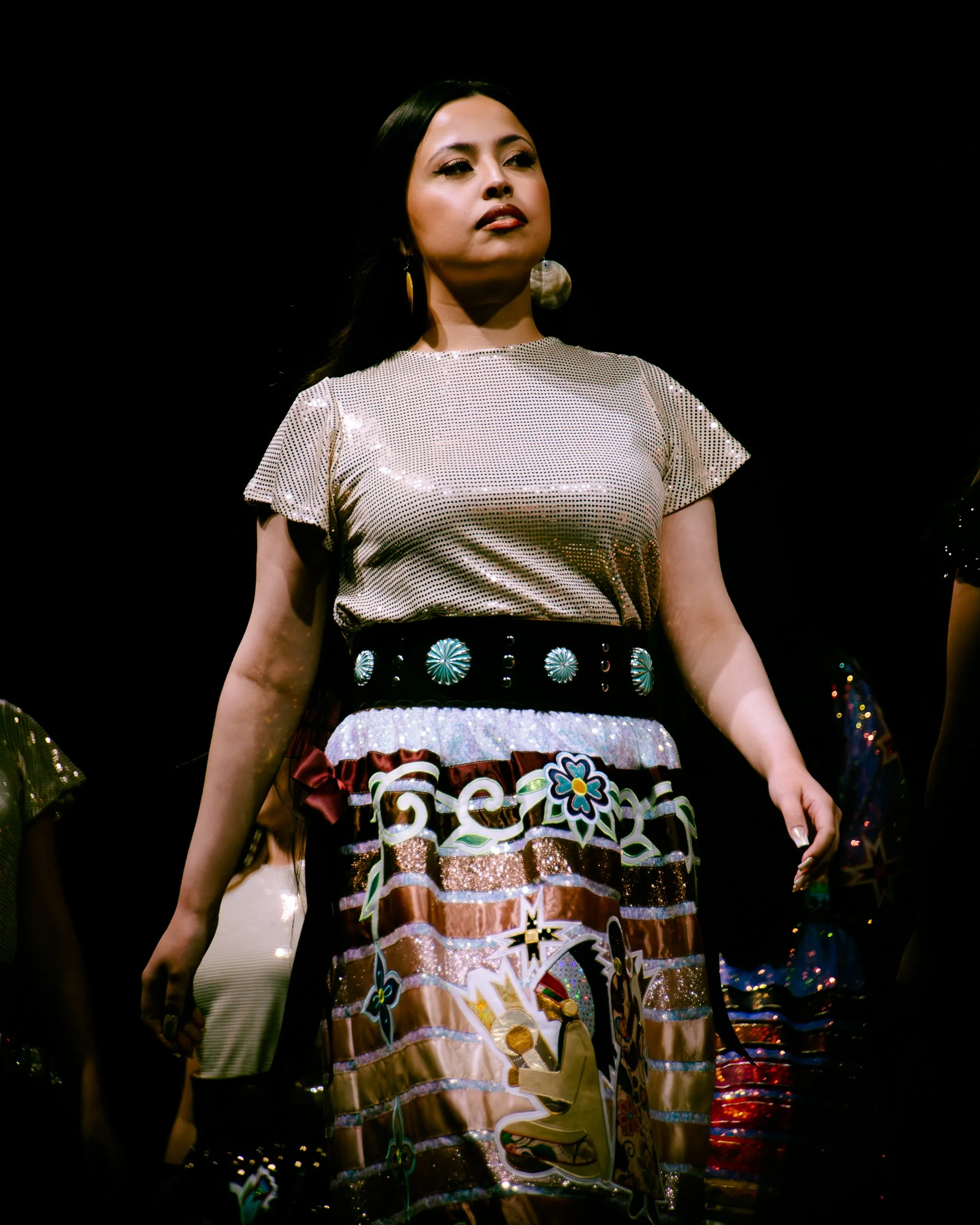 Fashion model on stage wearing a metallic top with a shiny skirt decorated with colorful patterns, including a stylized animal motif.