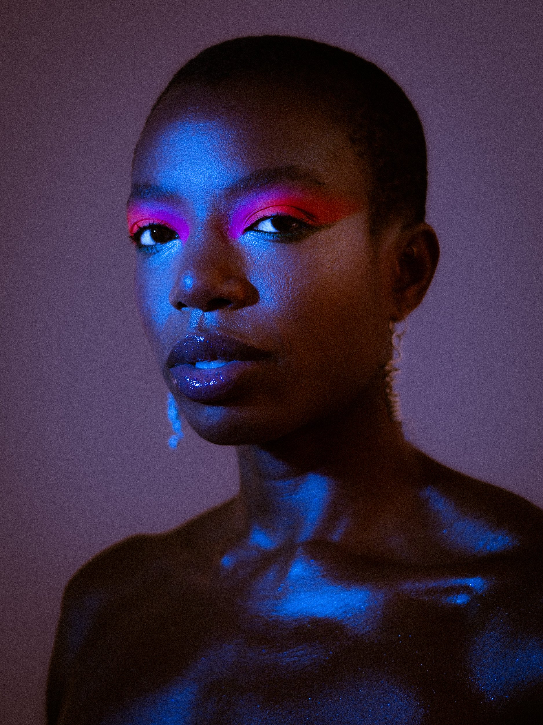 Close-up portrait of a Black woman model with short hair, neon pink and blue makeup, and shimmering black body paint, looking at the camera with a neutral expression against a gradient background, A woman wearing bright makeup, earrings, a rainbow be