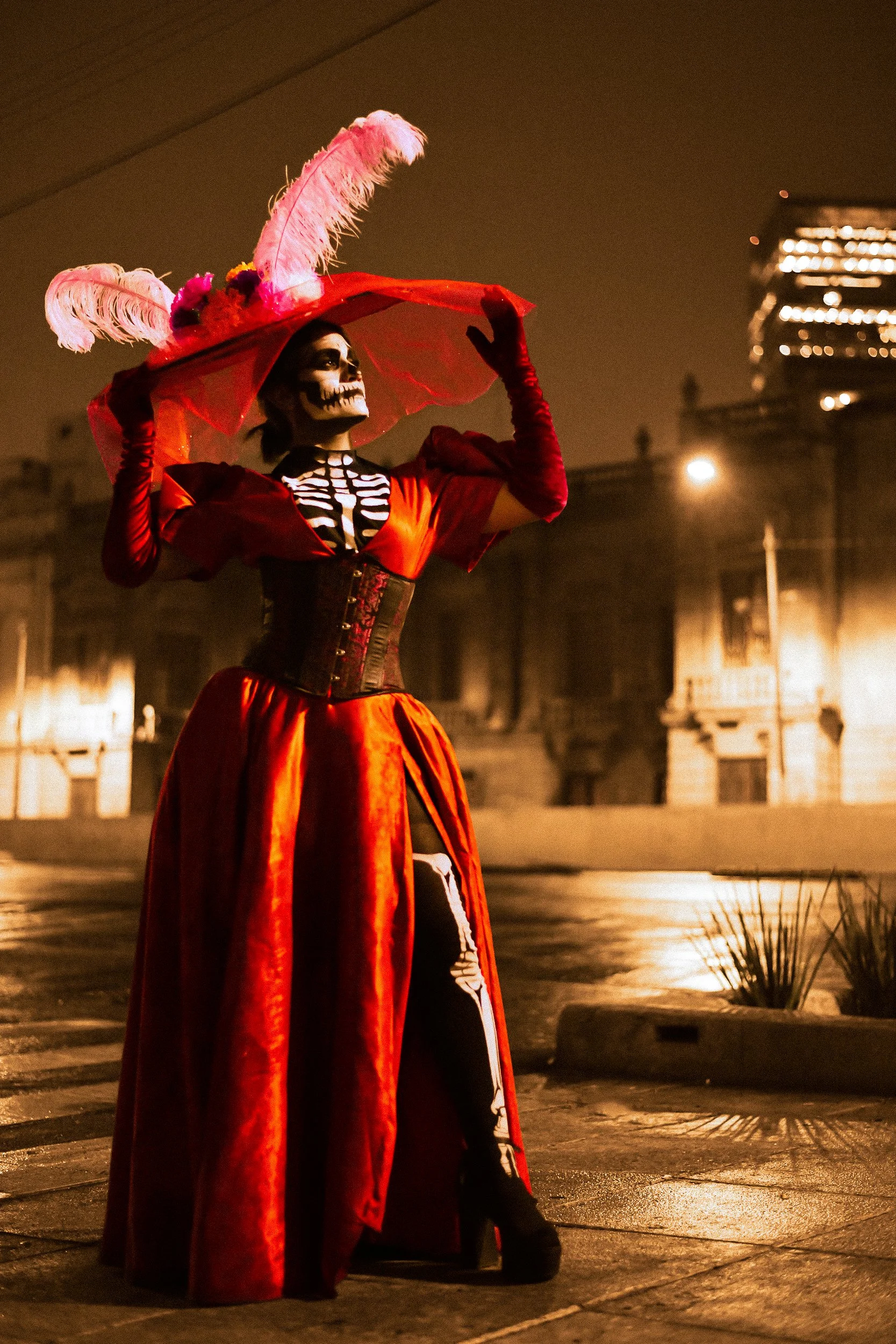 A person dressed in a skeleton costume with a red and black dress and a wide-brimmed hat decorated with pink feathers and flowers, standing outdoors at night with city buildings in the background.