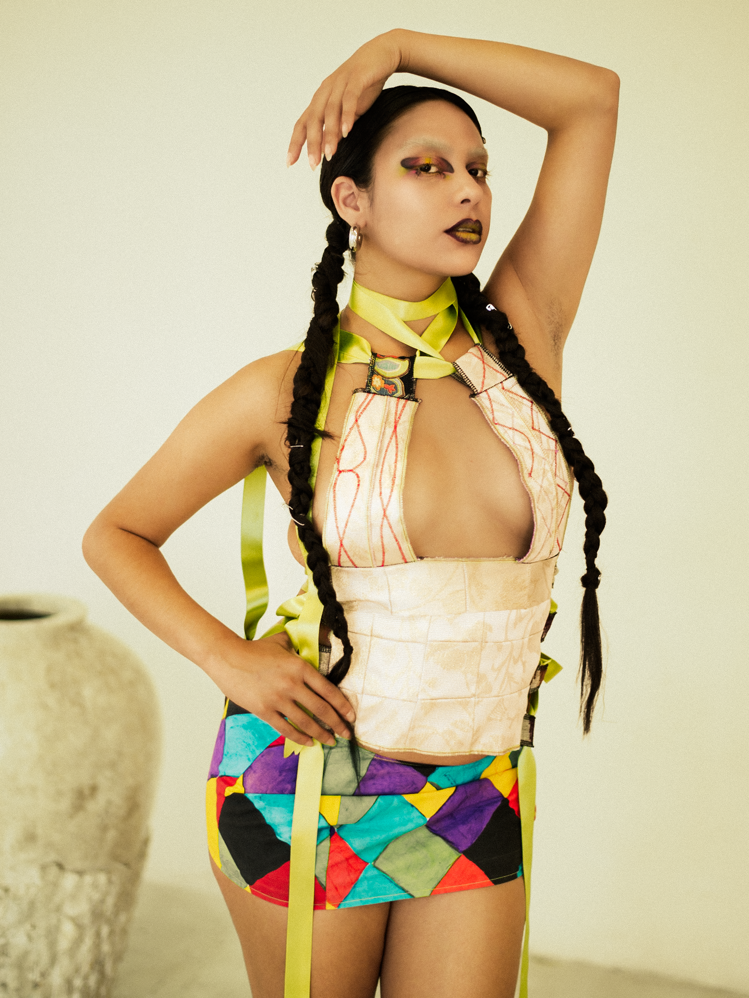 A woman model with long braided hair wearing multicolored outfit, yellow makeup, and white boots, posing indoors against a plain wall. photographed during a studio photography session in Los Angeles for a fashion editorial collaboration with fashion 