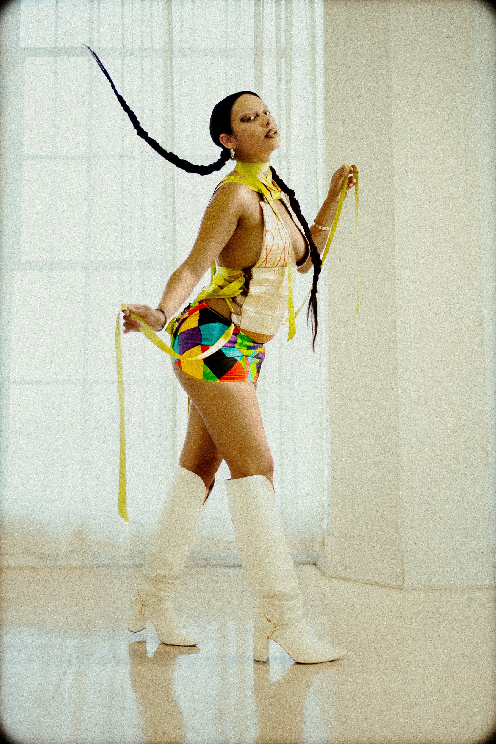 A woman in a colorful outfit with long braids, high white boots, and jewelry posing indoors near a window, photographed during a studio photography session in Los Angeles for a fashion editorial collaboration with fashion photographer