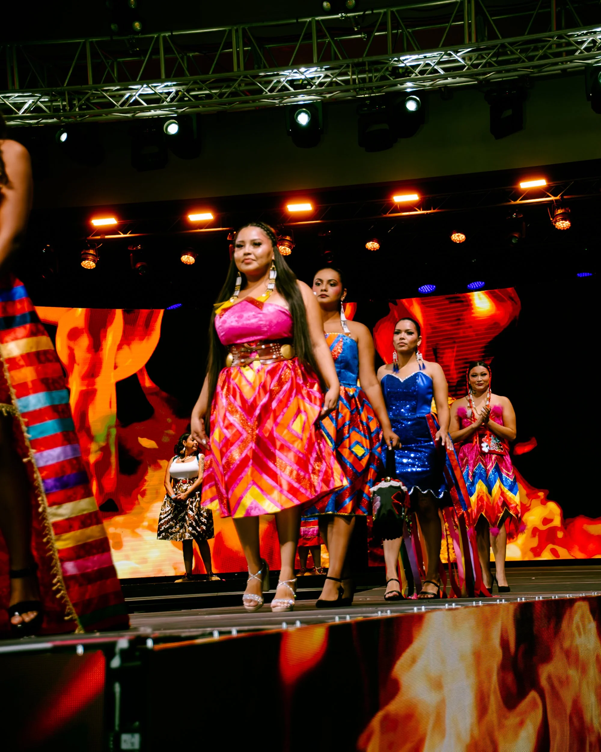Women in colorful traditional dresses walking on stage during a fashion show with flames and fiery background projections.