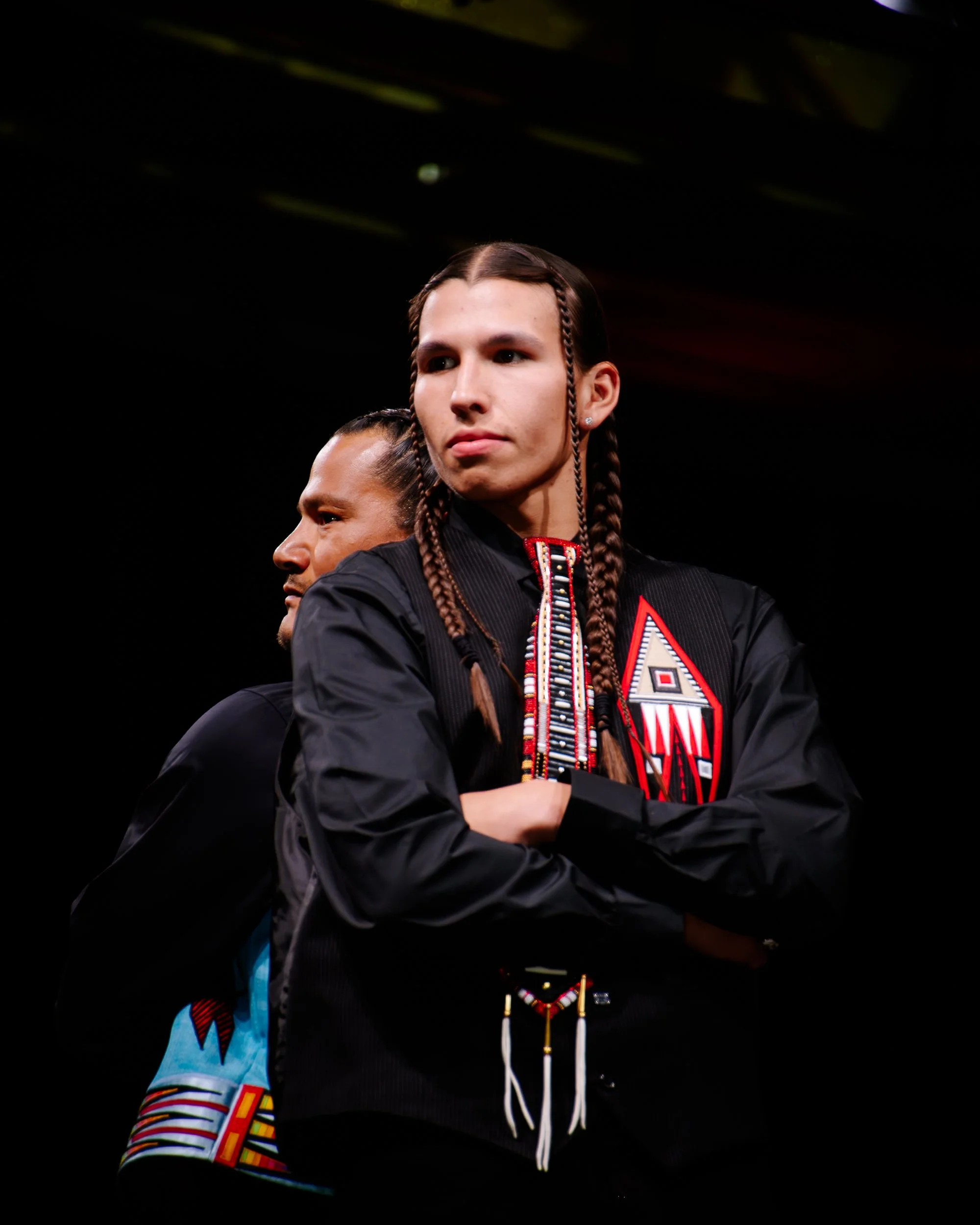A man with braided hair standing with arms crossed next to another man, both wearing black jackets with colorful designs.