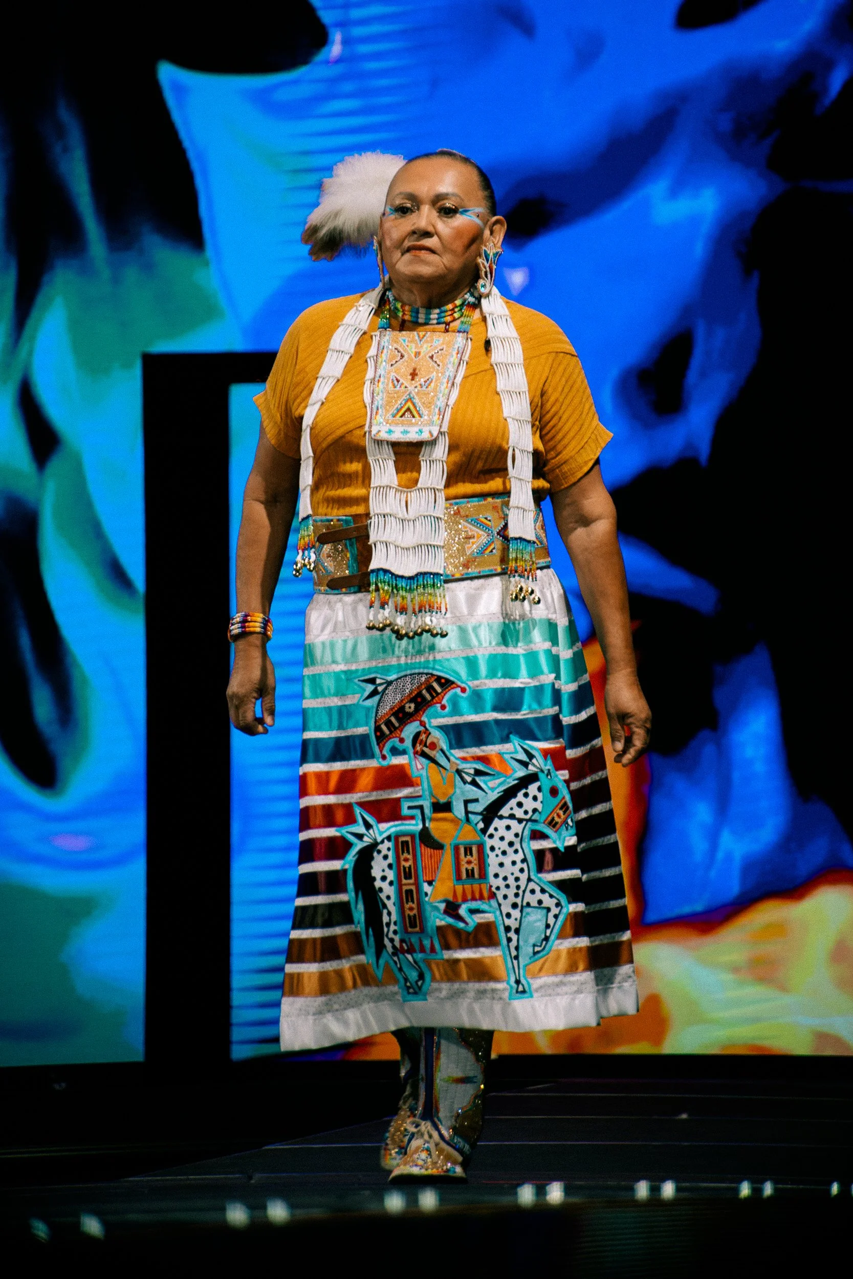 A woman walking on stage dressed in traditional Indigenous attire, including a patterned skirt, jewelry, and a feather in her hair, set against a colorful, artistic background.