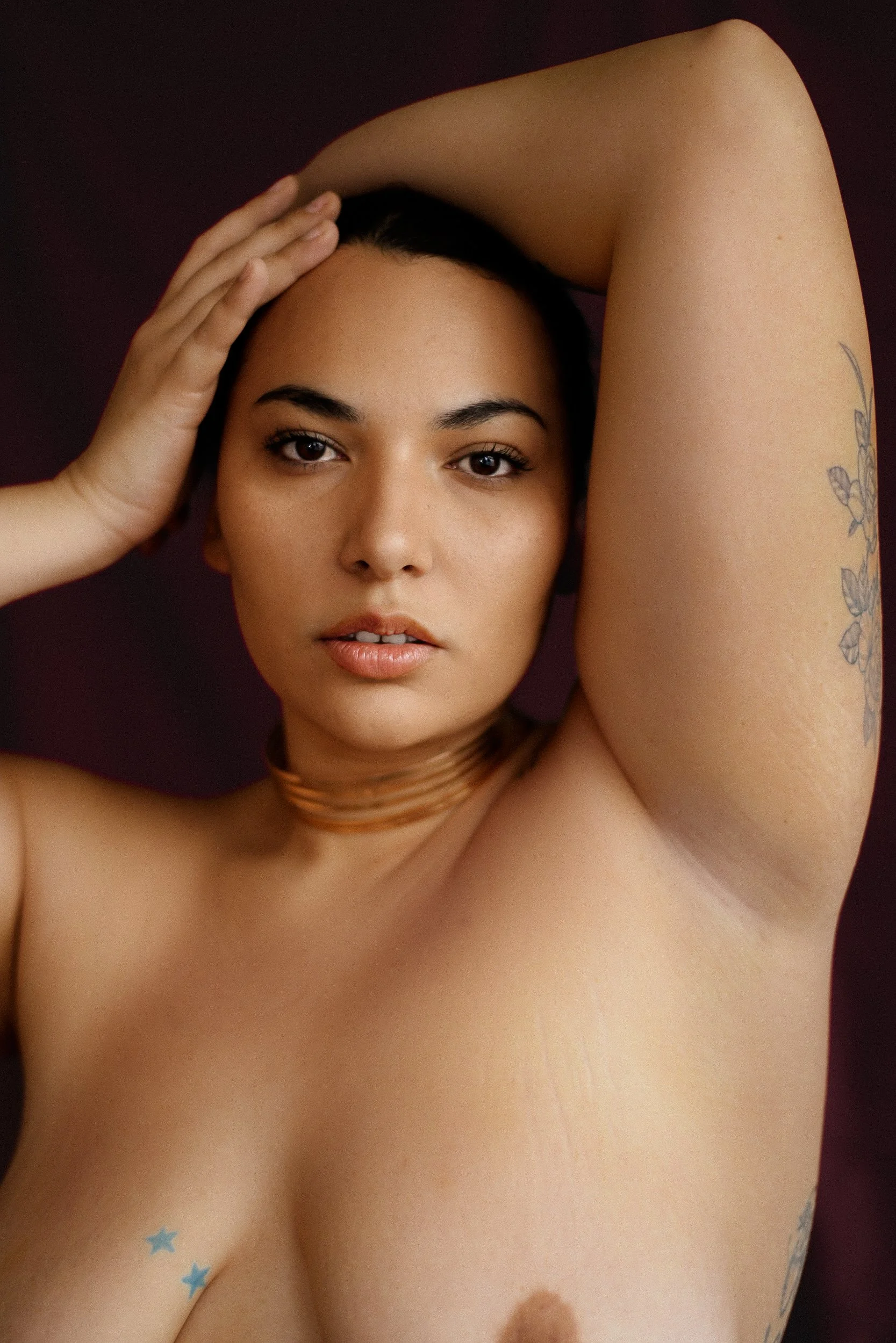 A topless woman with dark hair, tattoos on her arm and chest, and a choker, posing with her arm raised and hand on her head, looking directly at the camera against a dark background, photographed during a jewelry editorial photography session in Los 