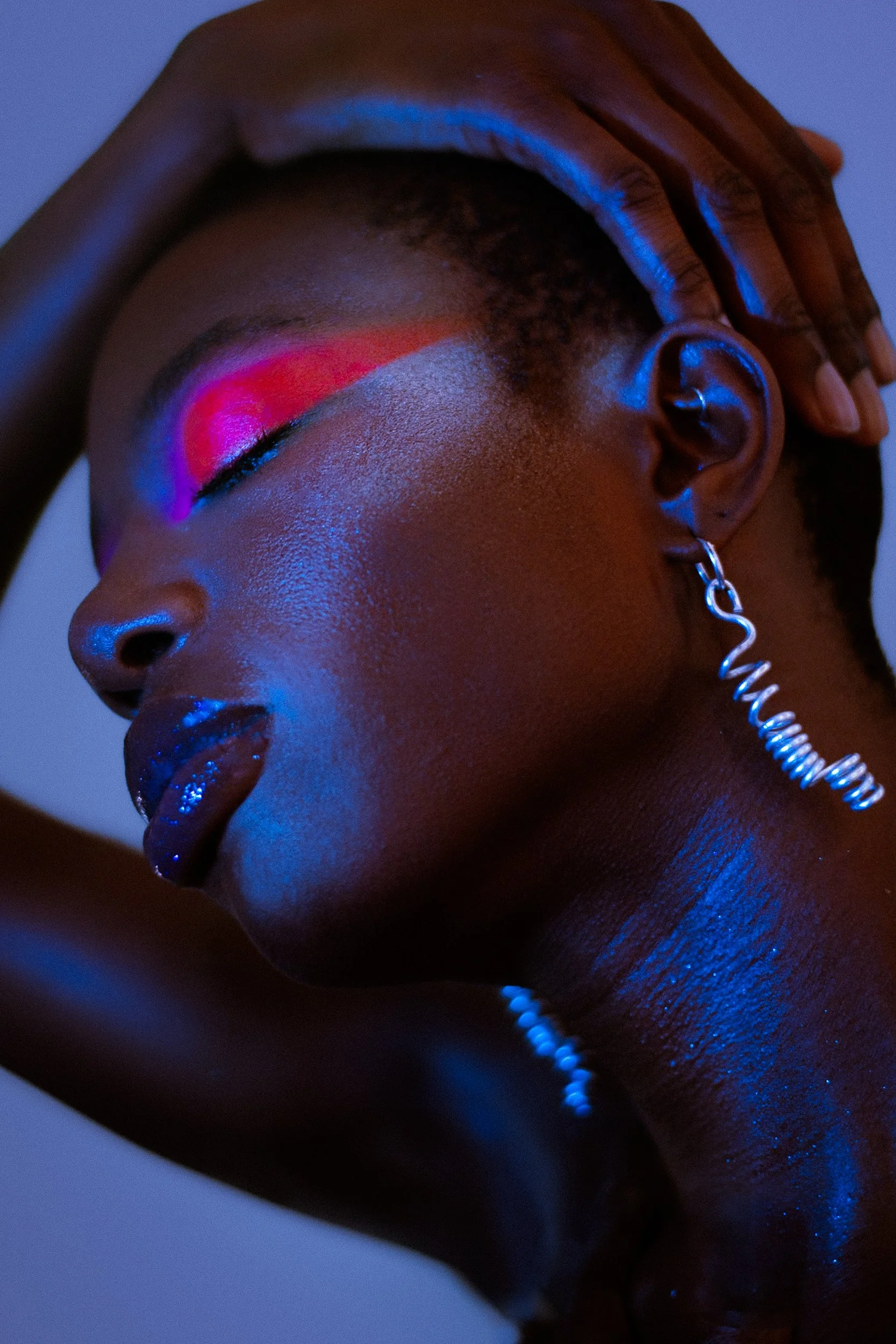 Close-up of a woman with rainbow-colored eye makeup and holographic lips, wearing spiral earrings and a pearl necklace, with her hand resting on her head. The lighting is blue and pink, shot for an editorial fashion photography session in Los Angeles