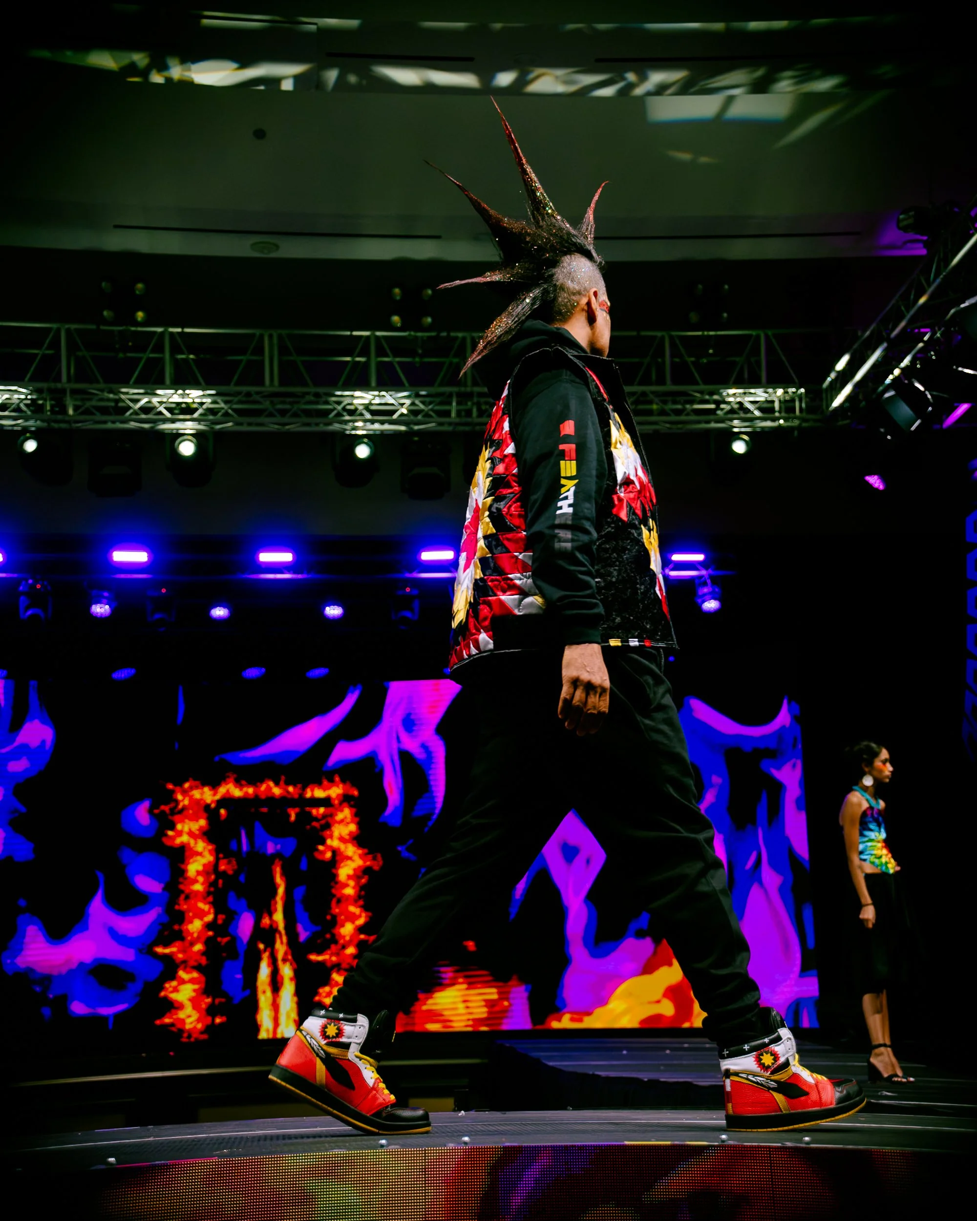 Person with a large, spiked mohawk hairstyle walking on a runway during a fashion show with colorful, animated background graphics.