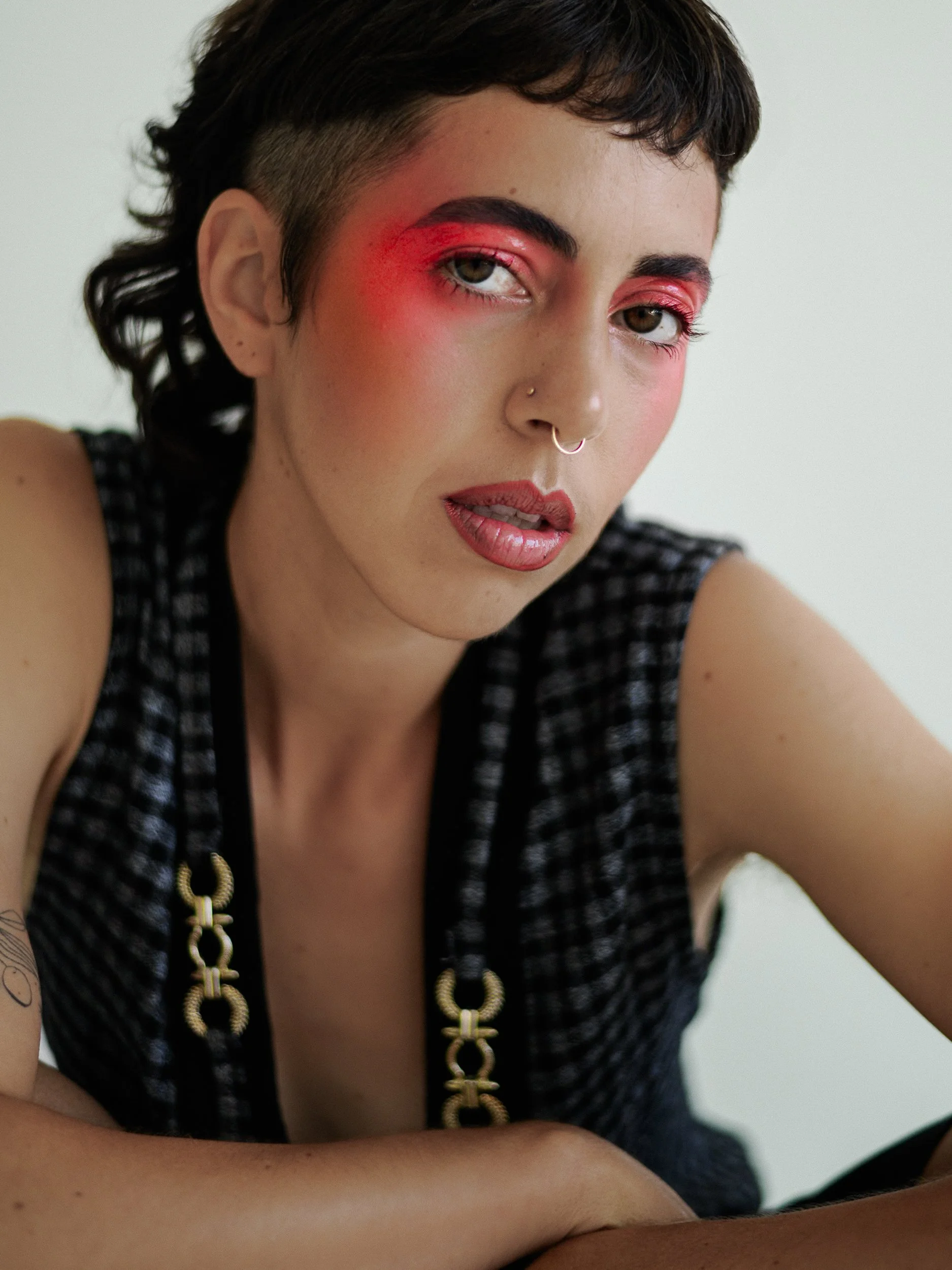 A person with short dark hair, bold makeup including pink eyeshadow and lipstick, a nose ring, and a tattoo on their left arm, wearing a sleeveless checkered top with gold chain details, posing against a plain white background, photographed during a 