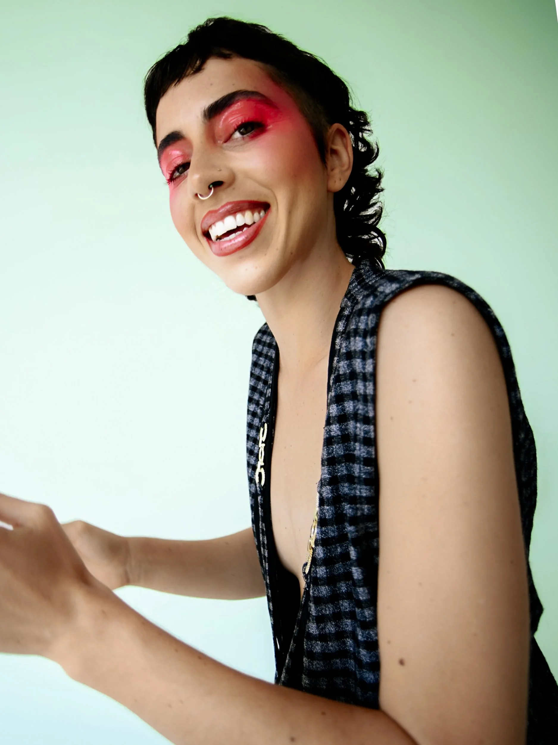 A person with short dark hair, pink eyeshadow, hoop septum piercing, wearing a sleeveless checkered top, smiling and looking at the camera against a plain background, photographed during a studio photography session in Los Angeles for a jewelry edito