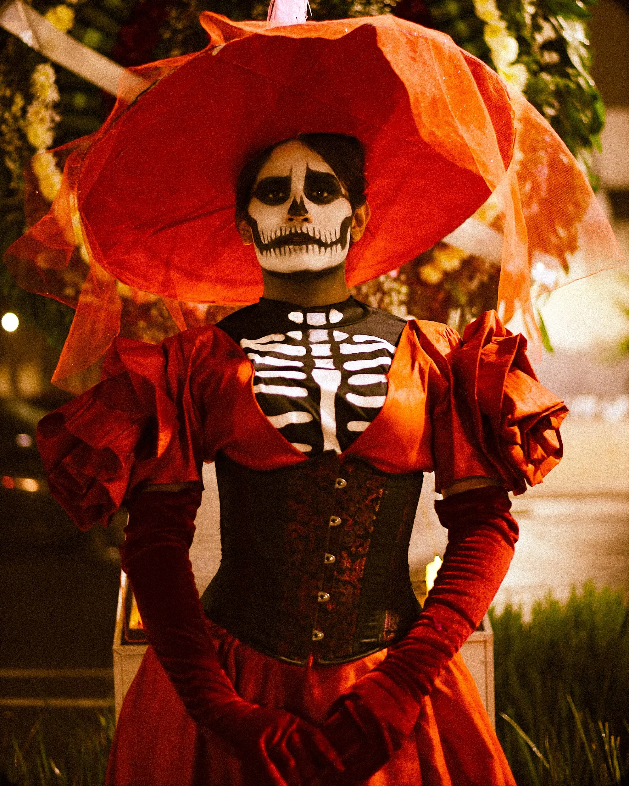 Person dressed in a skeleton costume with painted face, wearing a red wide-brimmed hat and red dress, standing outdoors at night with flowers in the background.