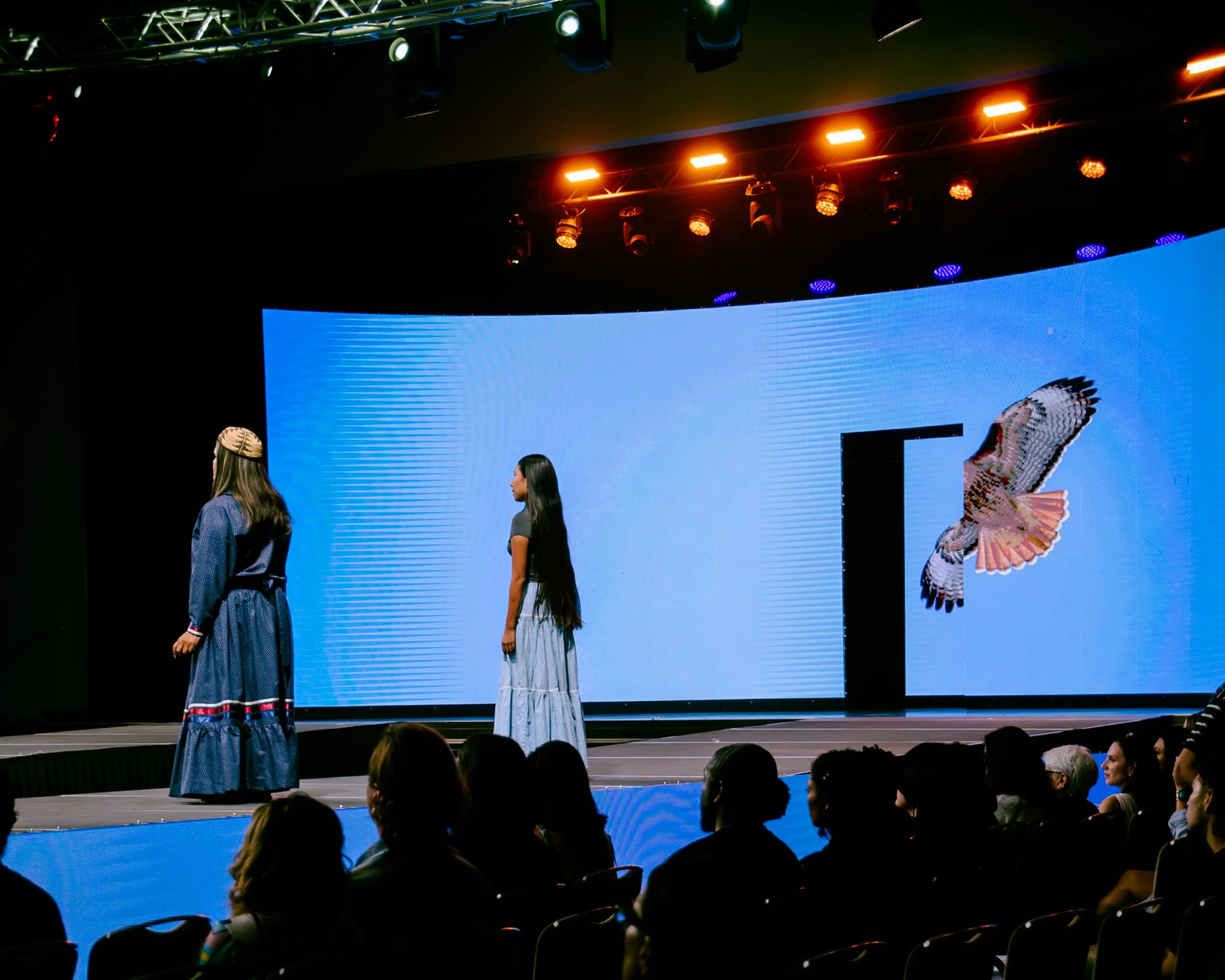 A fashion show with two models walking on stage, one in a long dress, and the other in a shorter dress, with a large digital screen behind them displaying a butterfly on a blue background, and an audience seated in front.