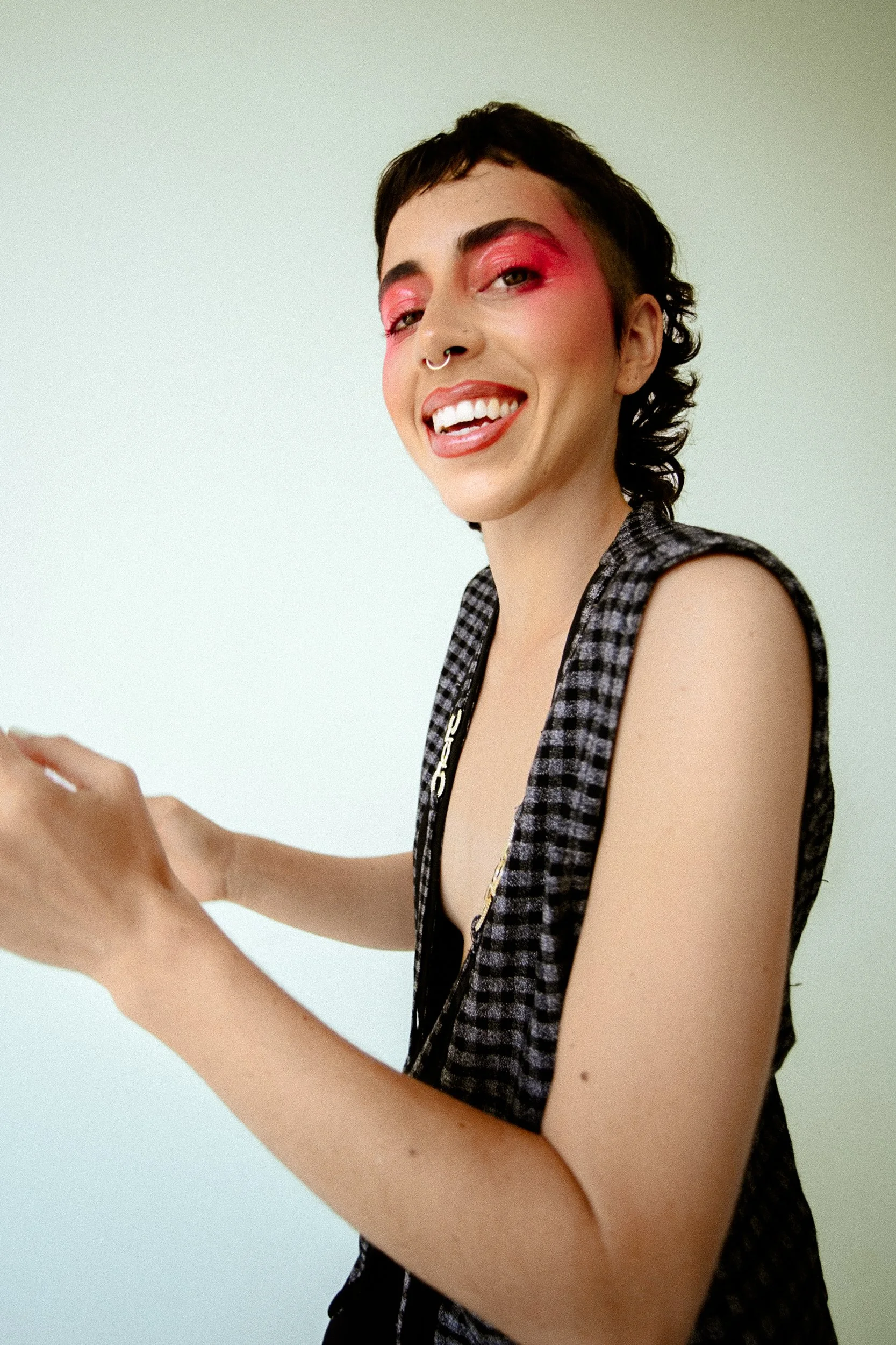 A person with short dark hair, pink eyeshadow, hoop septum piercing, wearing a sleeveless checkered top, smiling and looking at the camera against a plain background, photographed during a studio photography session in Los Angeles for a jewelry edito