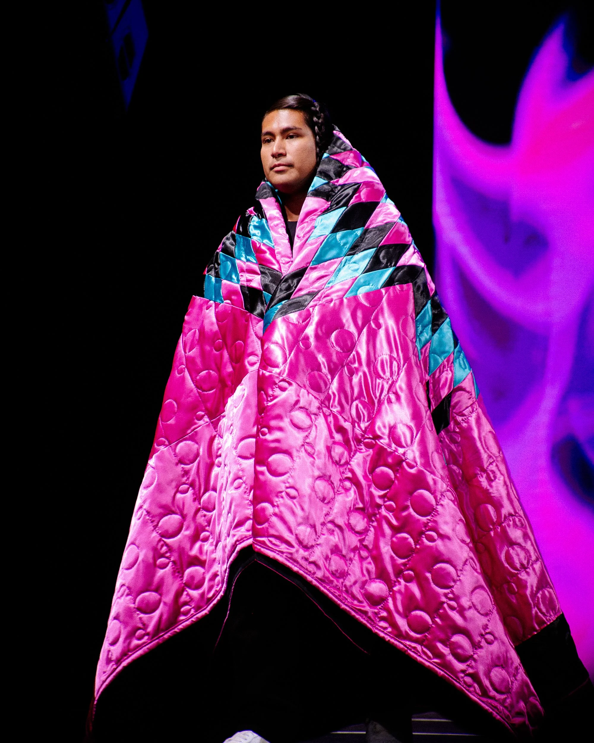 Model walking on runway in a large, colorful quilted pink, black, and blue cape with a patterned hood, against a dark background with purple lighting.