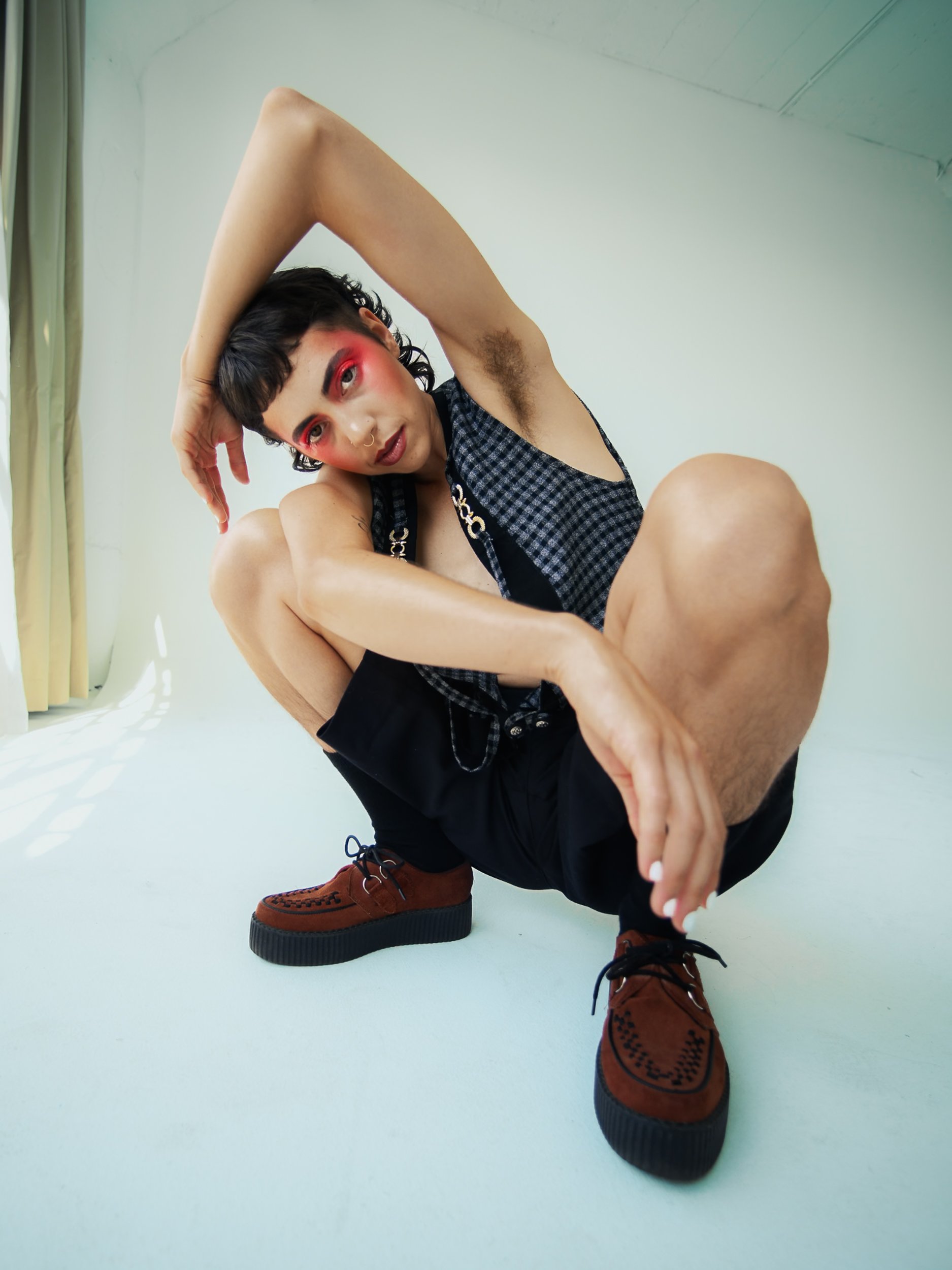 A person wearing a checkered black and blue sleeveless top, black shorts, and brown moccasin-style shoes poses on the floor with one arm over their head and the other hand on their foot, in a room with pale green walls and yellow curtains, photograph