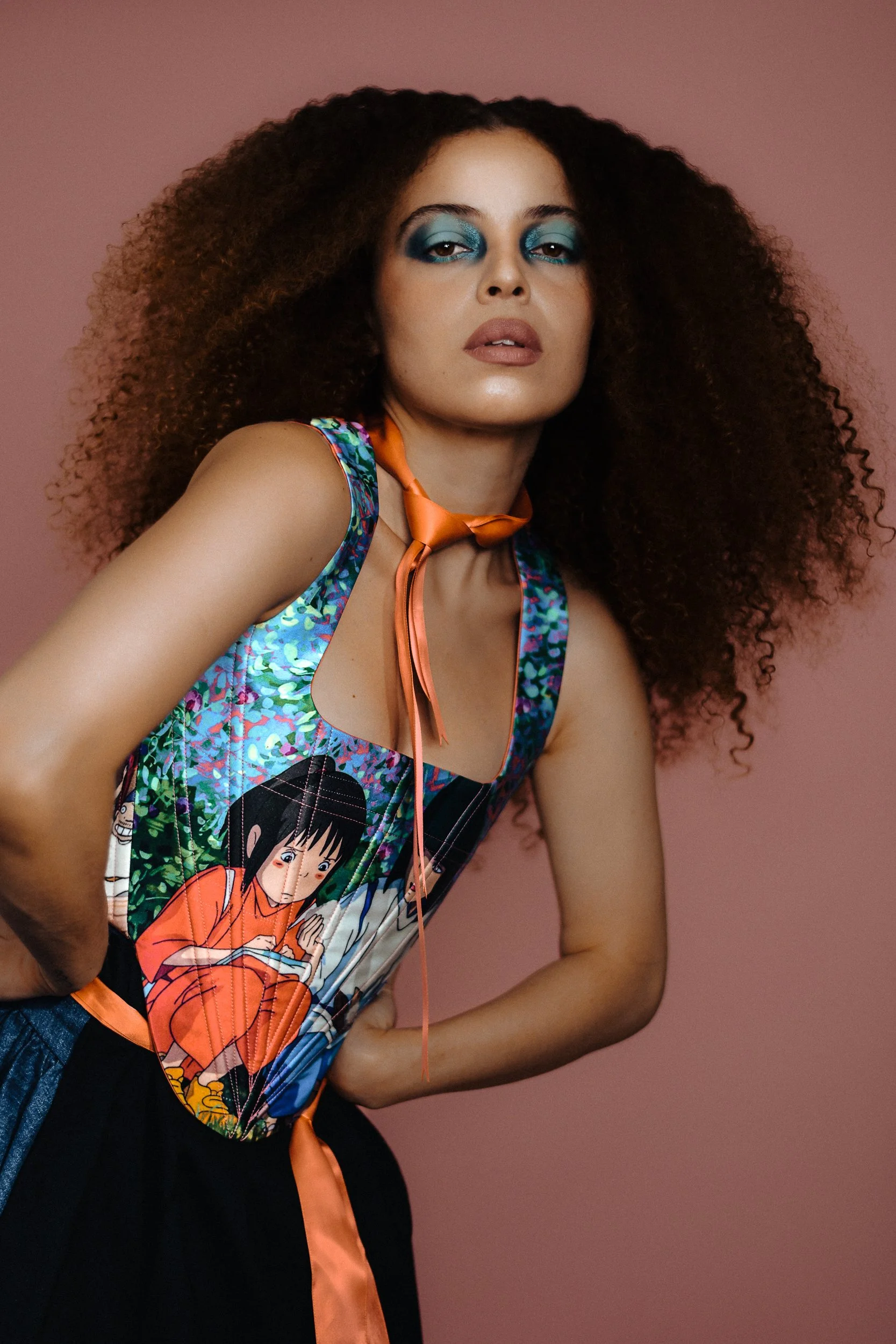 A woman with curly hair and bold blue eye makeup wearing a colorful sleeveless top with animated character print, an orange neck scarf, and black pants against a pink background, shot for an editorial fashion photography session in Los Angeles with f