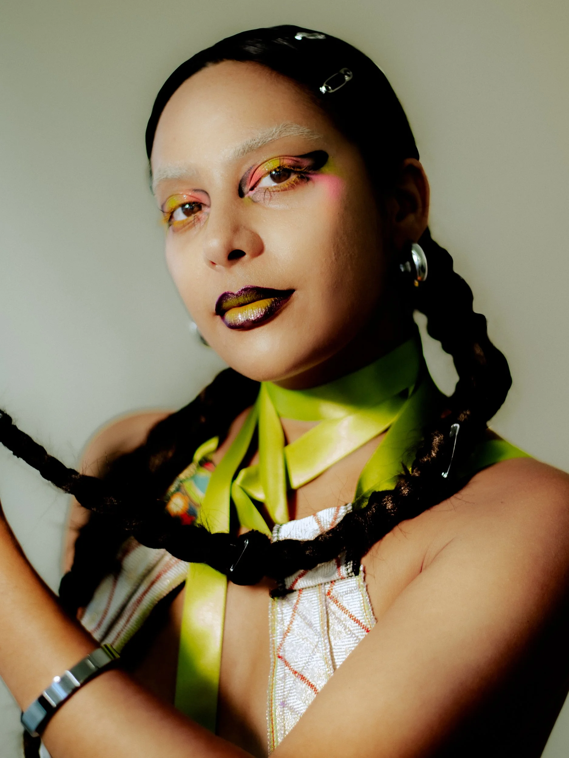 A person with bold, colorful makeup, black braided hair with clips, wearing a shiny lime green ribbon around the neck, earrings, and a patterned top, photographed during a fashion editorial photography session in Los Angeles with fashion photographer