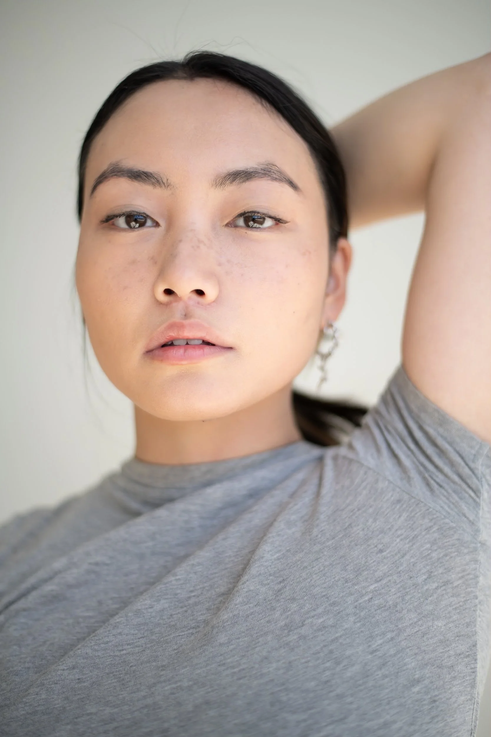 Close-up of a young woman with dark hair, light skin with freckles, and wearing a gray shirt, posing with her arm behind her head, photographed during a portrait photography session in Los Angeles with fashion photographer Gí Tenamá.