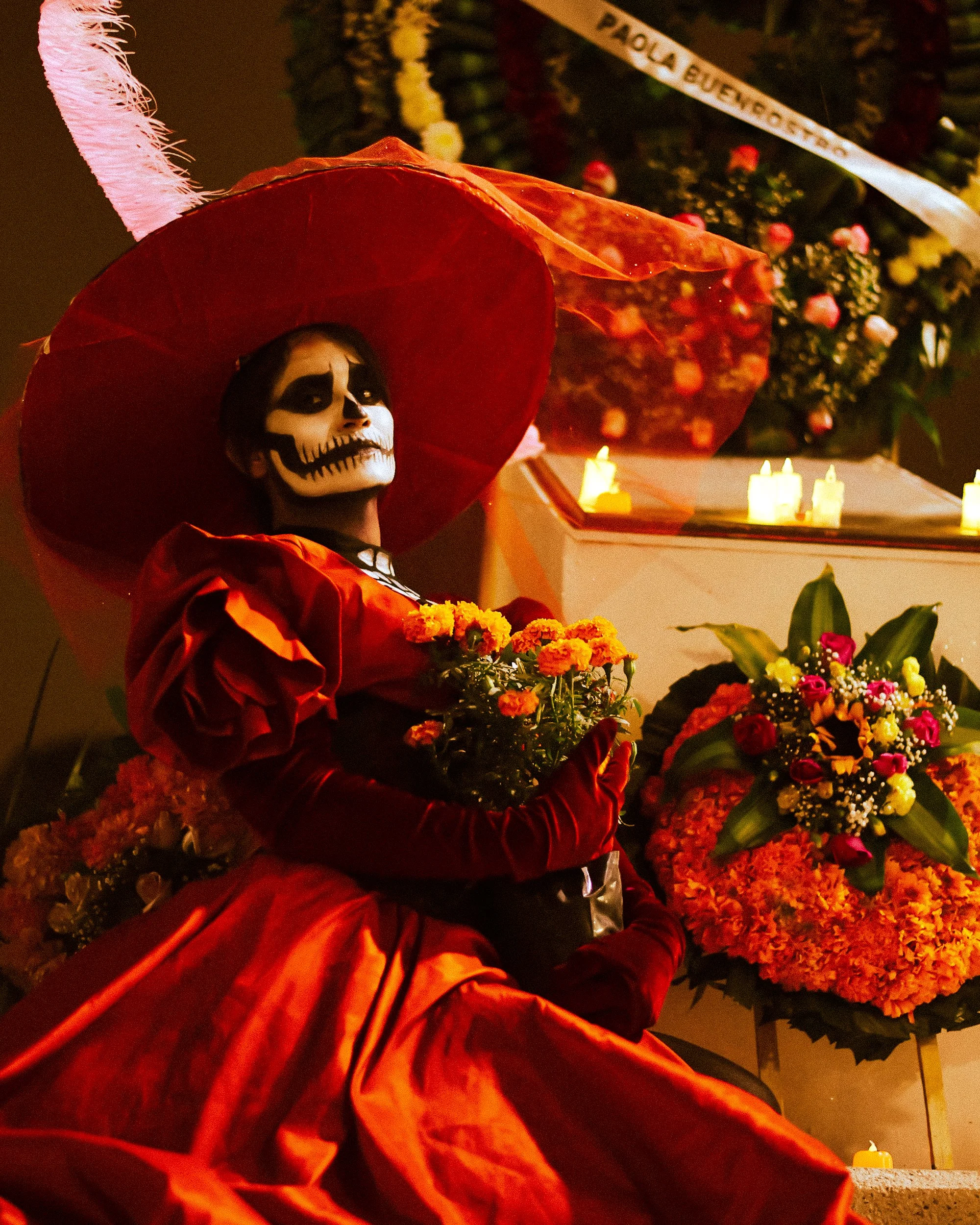 A person dressed in traditional Mexican attire with a skeleton face paint, holding flowers, sitting among colorful floral arrangements and candles, likely during the Day of the Dead celebration.