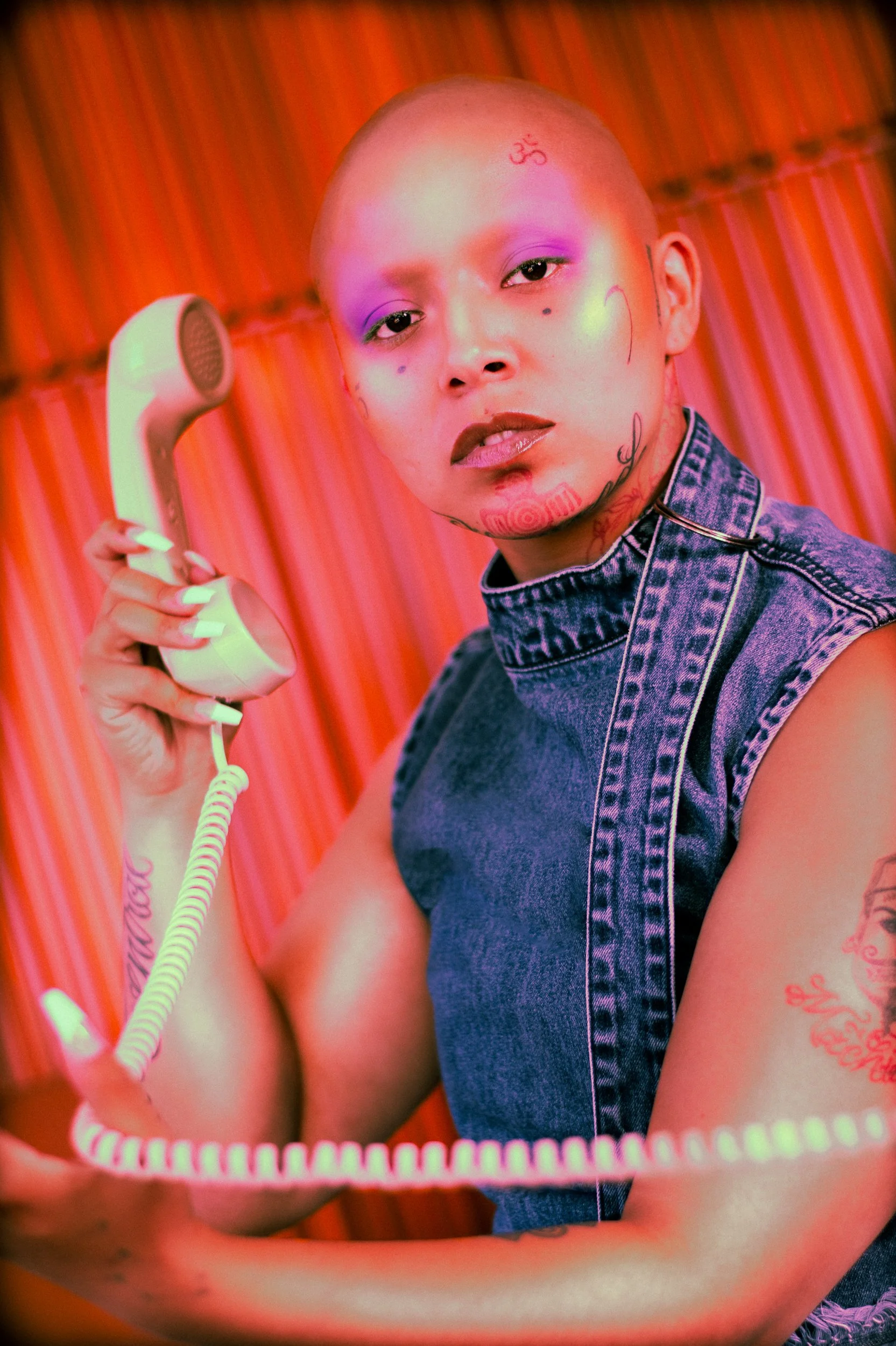 A person with tattoos and makeup dressed in a sleeveless denim top, holding an old-fashioned telephone receiver against a vibrant, striped background, photographed during a studio photography session in Los Angeles for a jewelry editorial collaborati