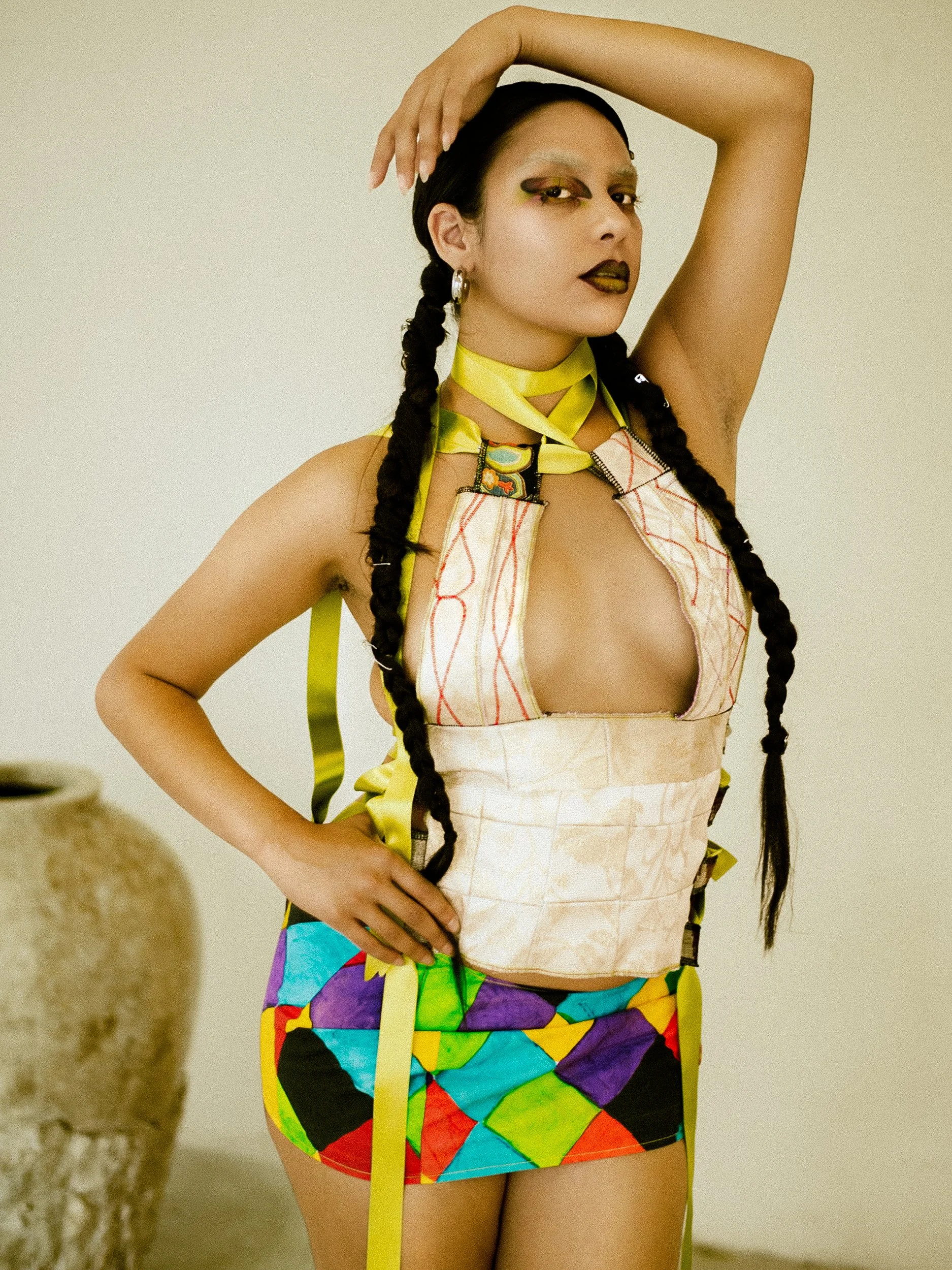 Woman with long braided hair and bold makeup, wearing a colorful patchwork skirt, a transparent top with red stitching, and a yellow ribbon around her neck, striking a pose with one hand on her hip and the other behind her head, photographed during a