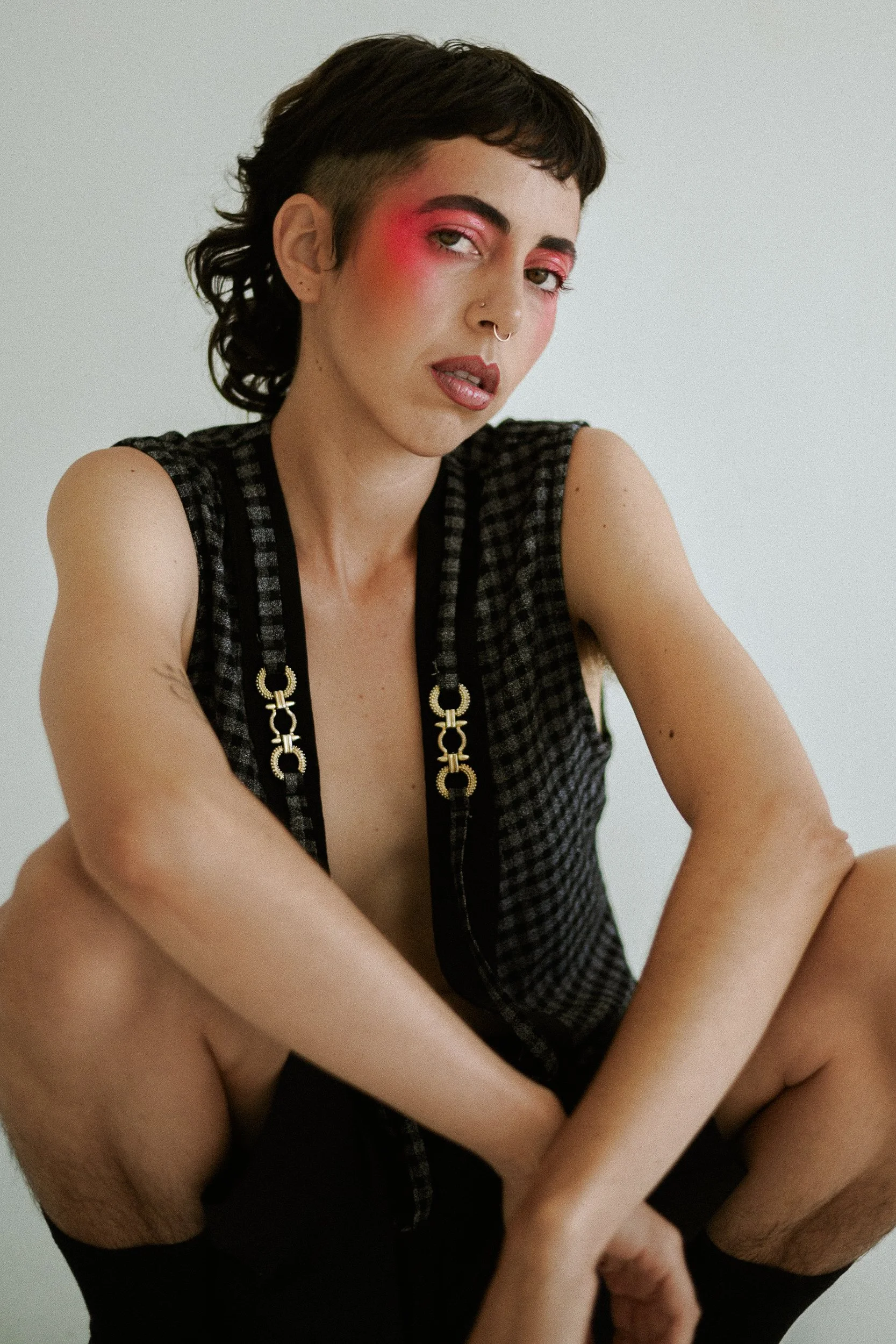 A person with short dark curly hair and pink eyeshadow, wearing a sleeveless black checkered top with gold chain details, sitting cross-legged against a plain white background, photographed during a studio photography session in Los Angeles for a fas
