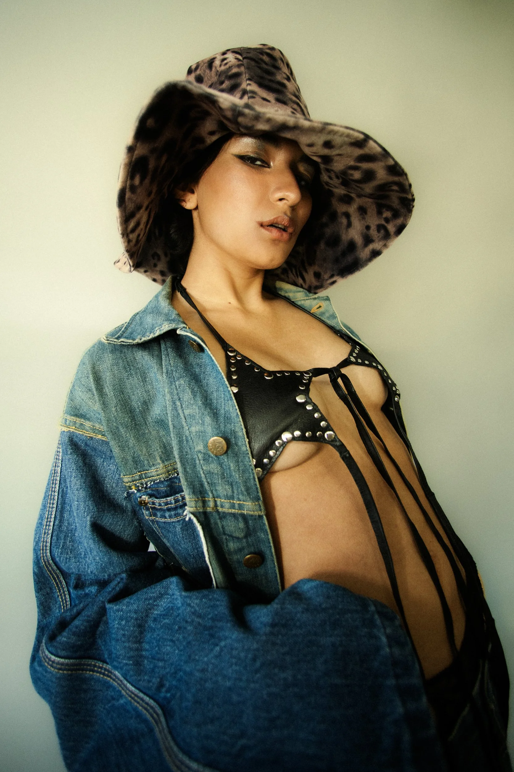 Woman wearing a large leopard-print hat, denim jacket, and a black studded leather top, posing against a plain background, photographed during a fashion editorial photography session in Los Angeles with fashion photographer Gí Tenamá..