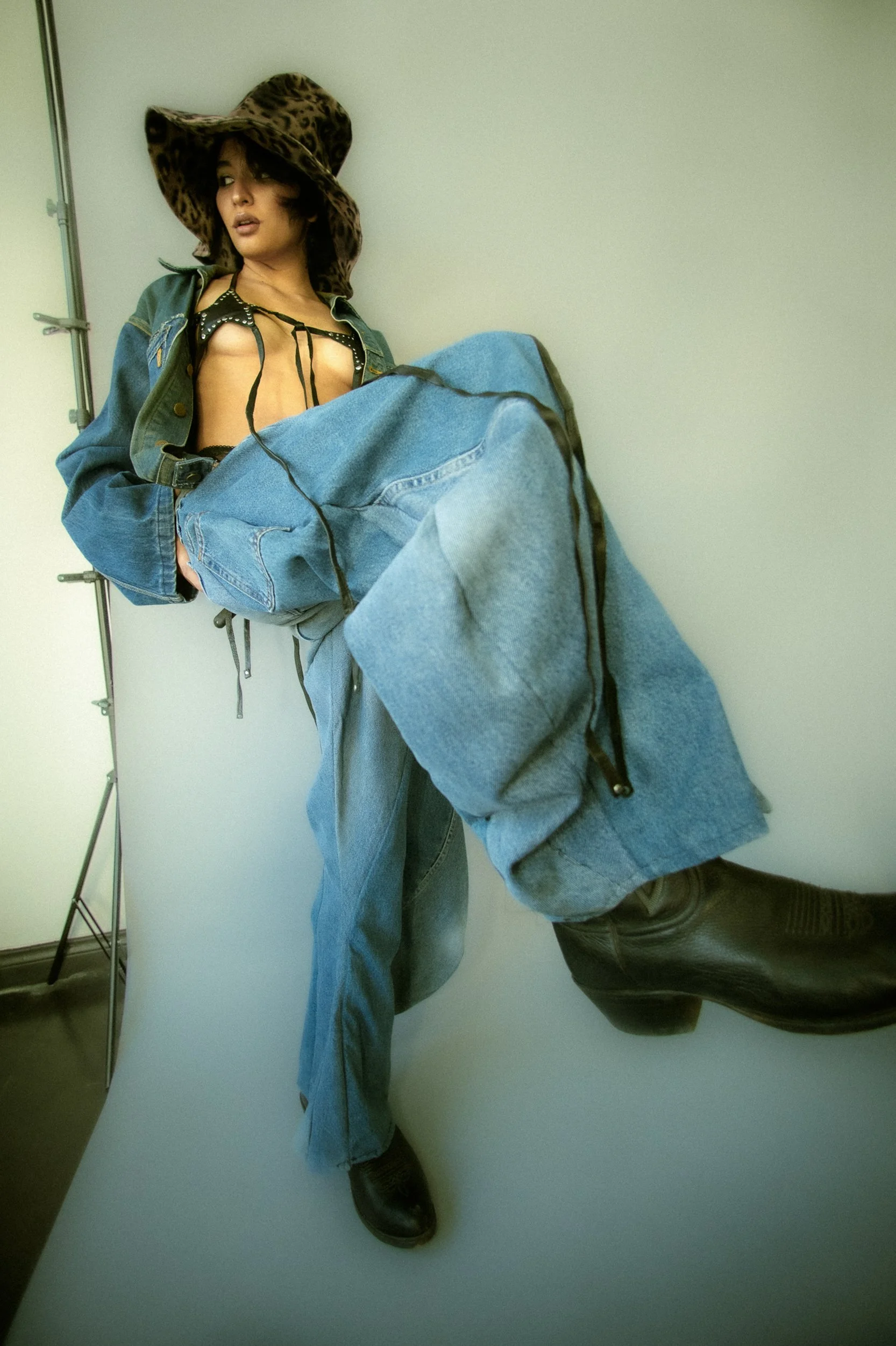 A woman with dark hair wearing a leopard-print wide-brimmed hat, a denim jacket, and oversized jeans pose against a pale backdrop, with one leg bent and her gaze directed away, photographed during a fashion editorial photography session in Los Angele