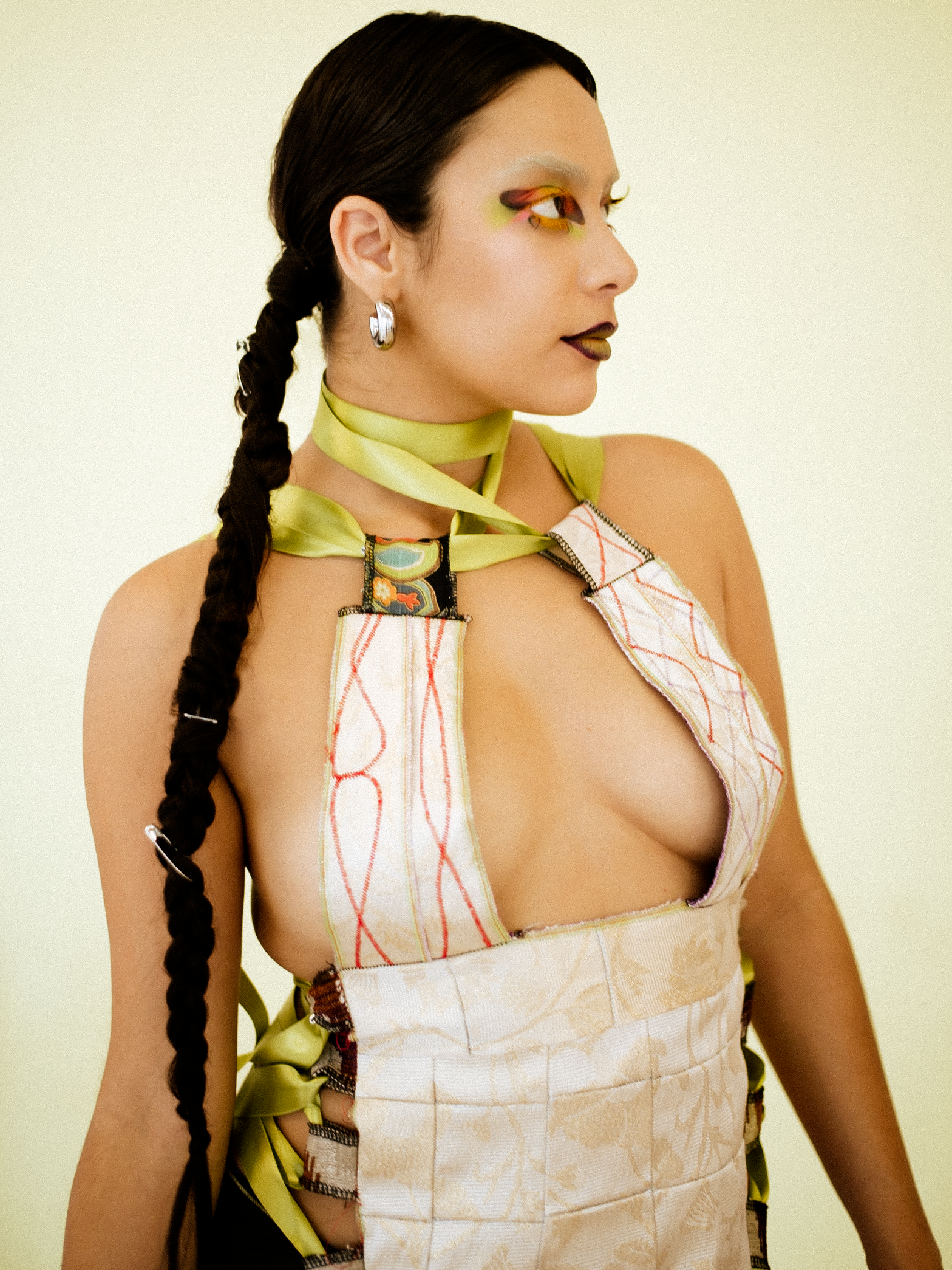A woman model with long braided hair wearing multicolored outfit, yellow makeup, and white boots, posing indoors against a plain wall. photographed during a studio photography session in Los Angeles for a fashion editorial collaboration with fashion 