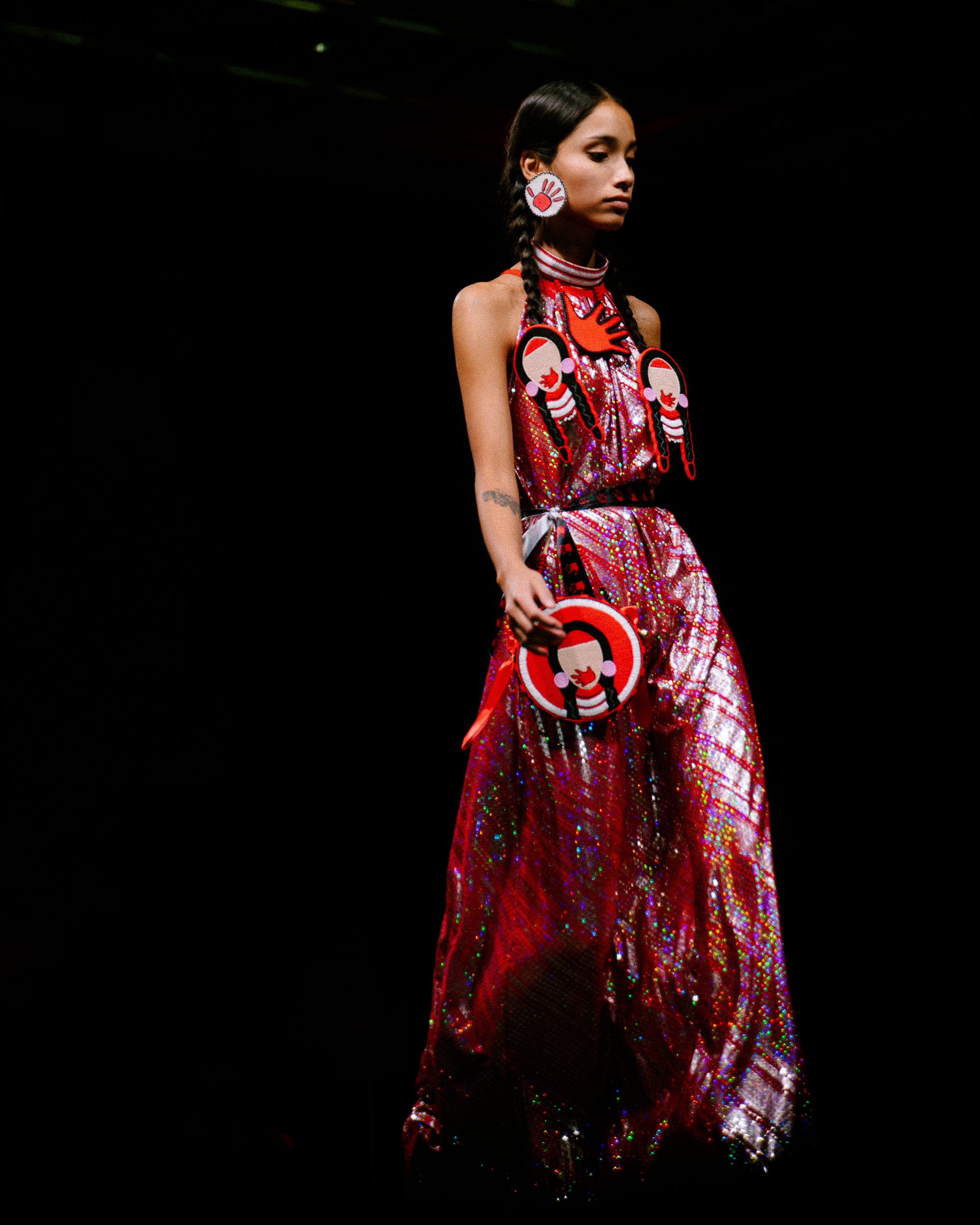 Fashion model walking on runway in colorful, shiny dress with bold, graphic jewelry and accessories against a black background.