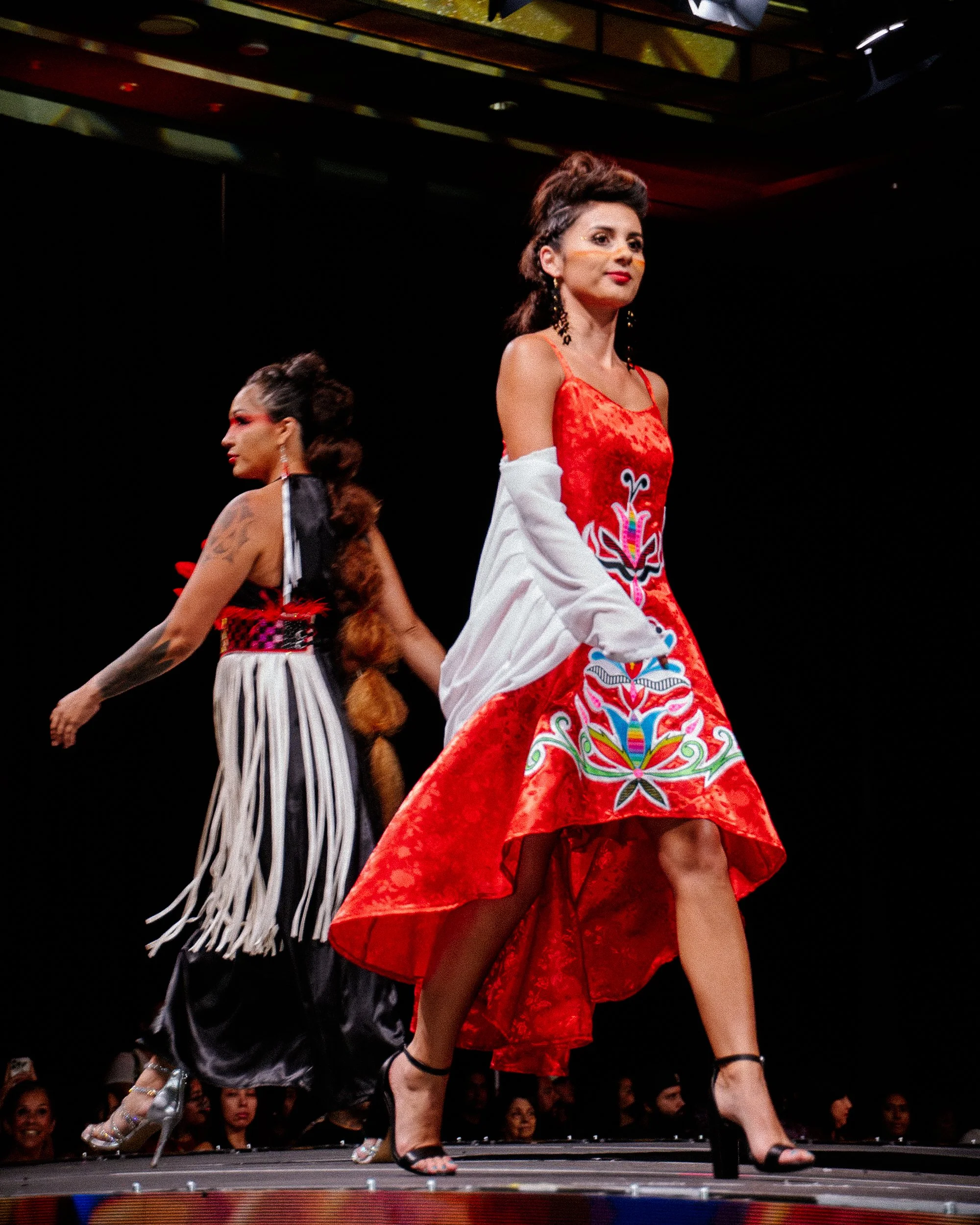 Two women walking on a runway, wearing colorful, elaborate dresses with bold patterns, and high heels, during a fashion show.