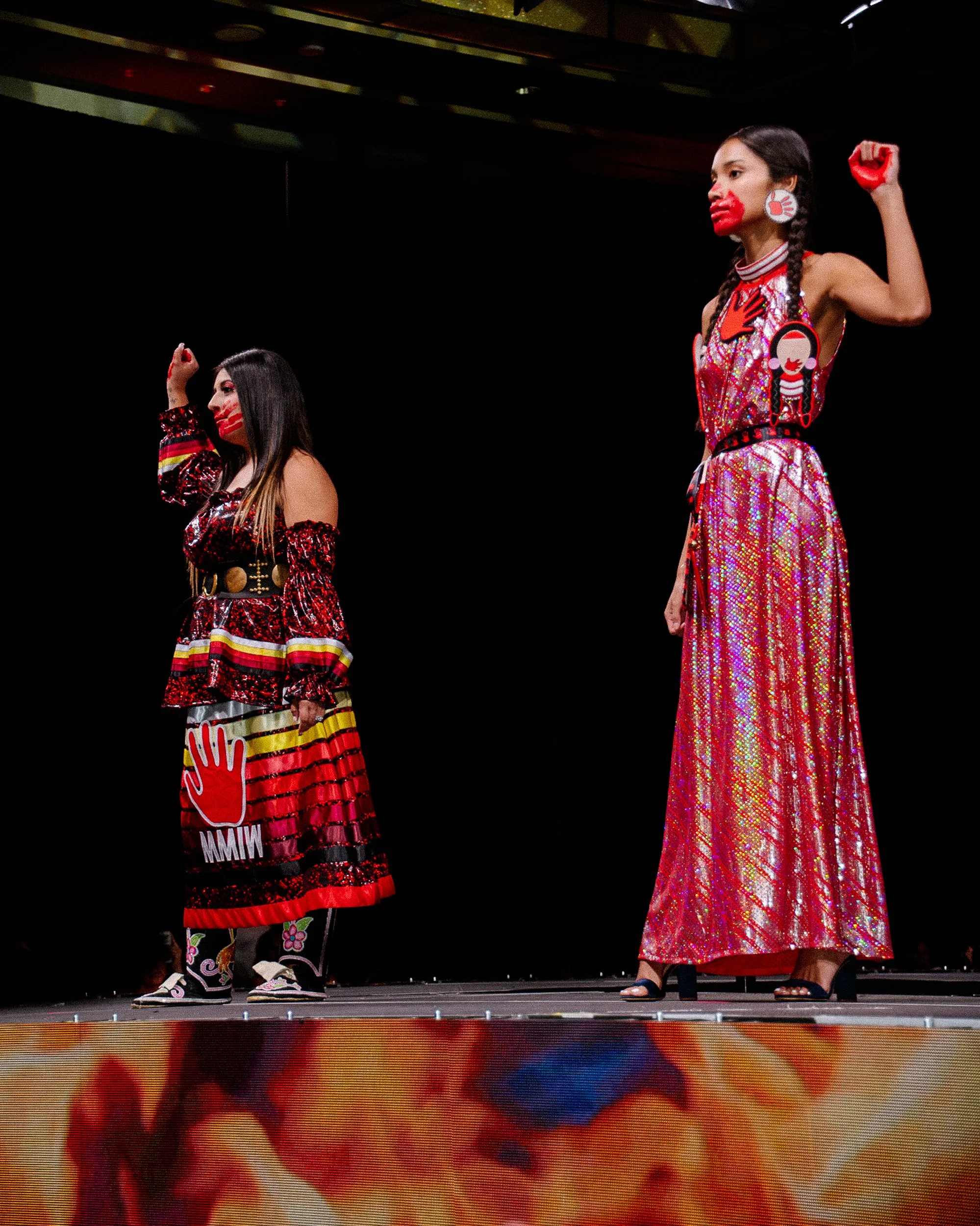 Two women on a stage wearing colorful, shiny outfits with face paint and symbolic jewelry, standing against a black backdrop.