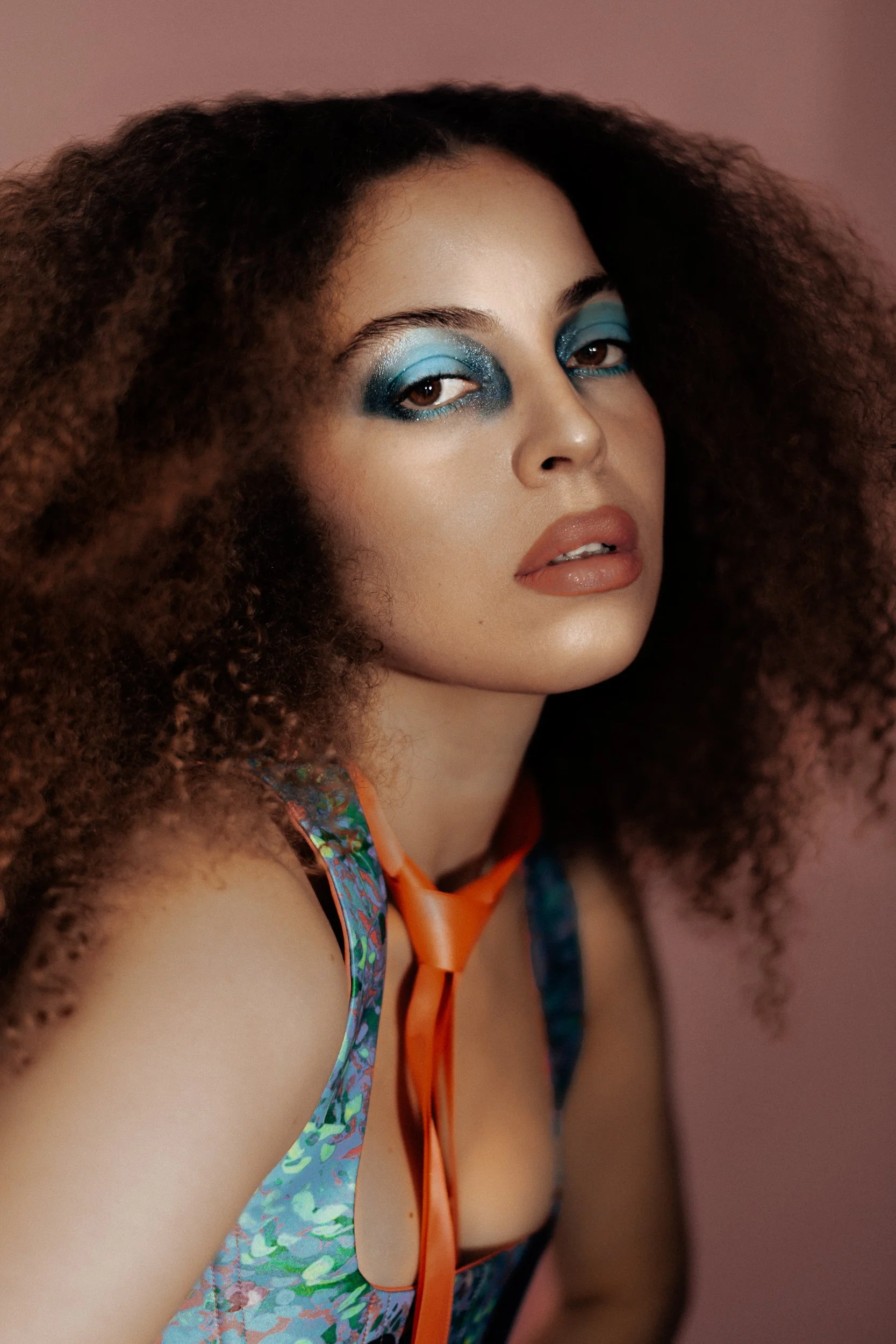 A woman with curly hair and blue eye makeup, wearing a colorful top with an orange ribbon, posing against a pink background, photographed during a fashion editorial photography session in Los Angeles with fashion photographer Gí Tenamá..