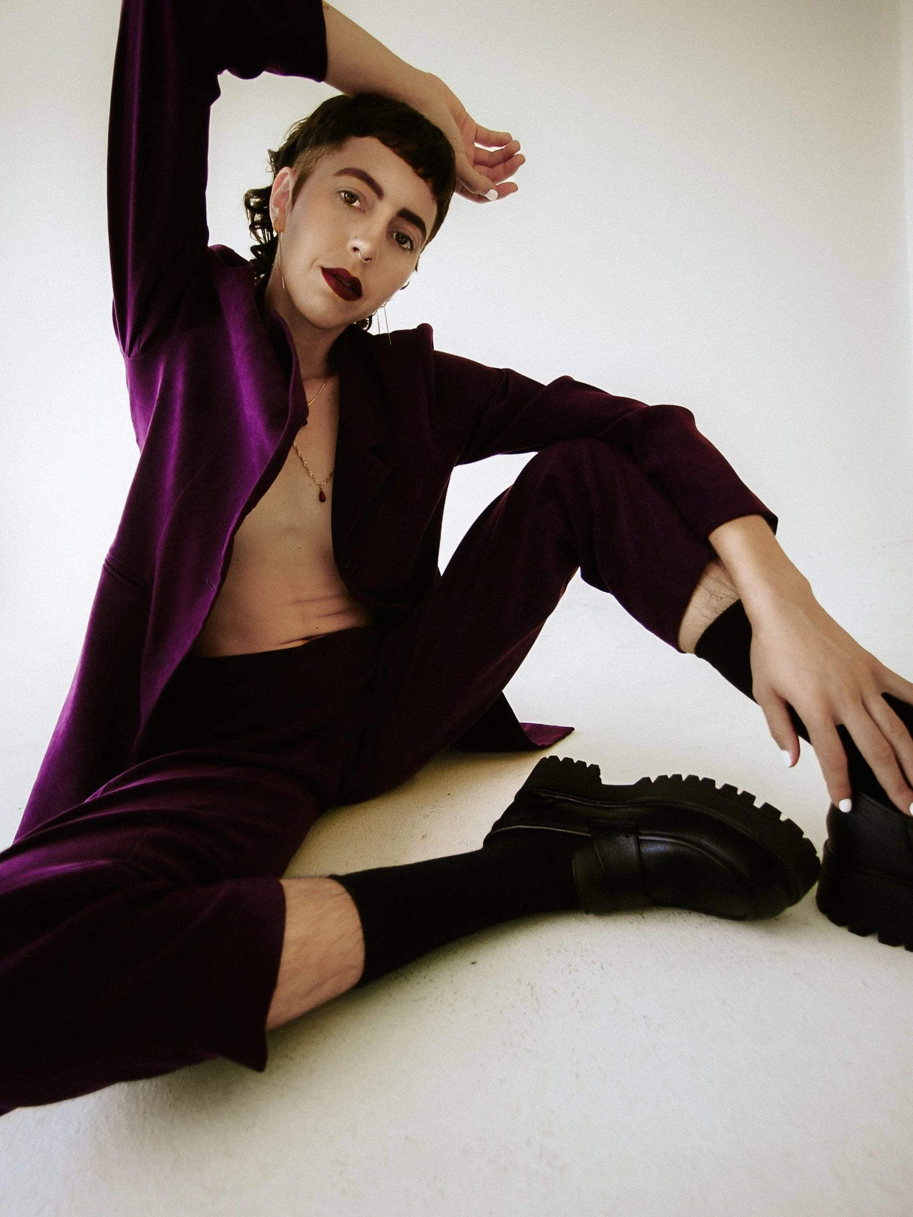 Close-up portrait of a queer nonbinary person with short, dark hair, purple eyebrows, and dark purple lipstick, wearing jewelry including earrings and necklaces, dressed in a dark purple shirt, against a plain white background, photographed during a 