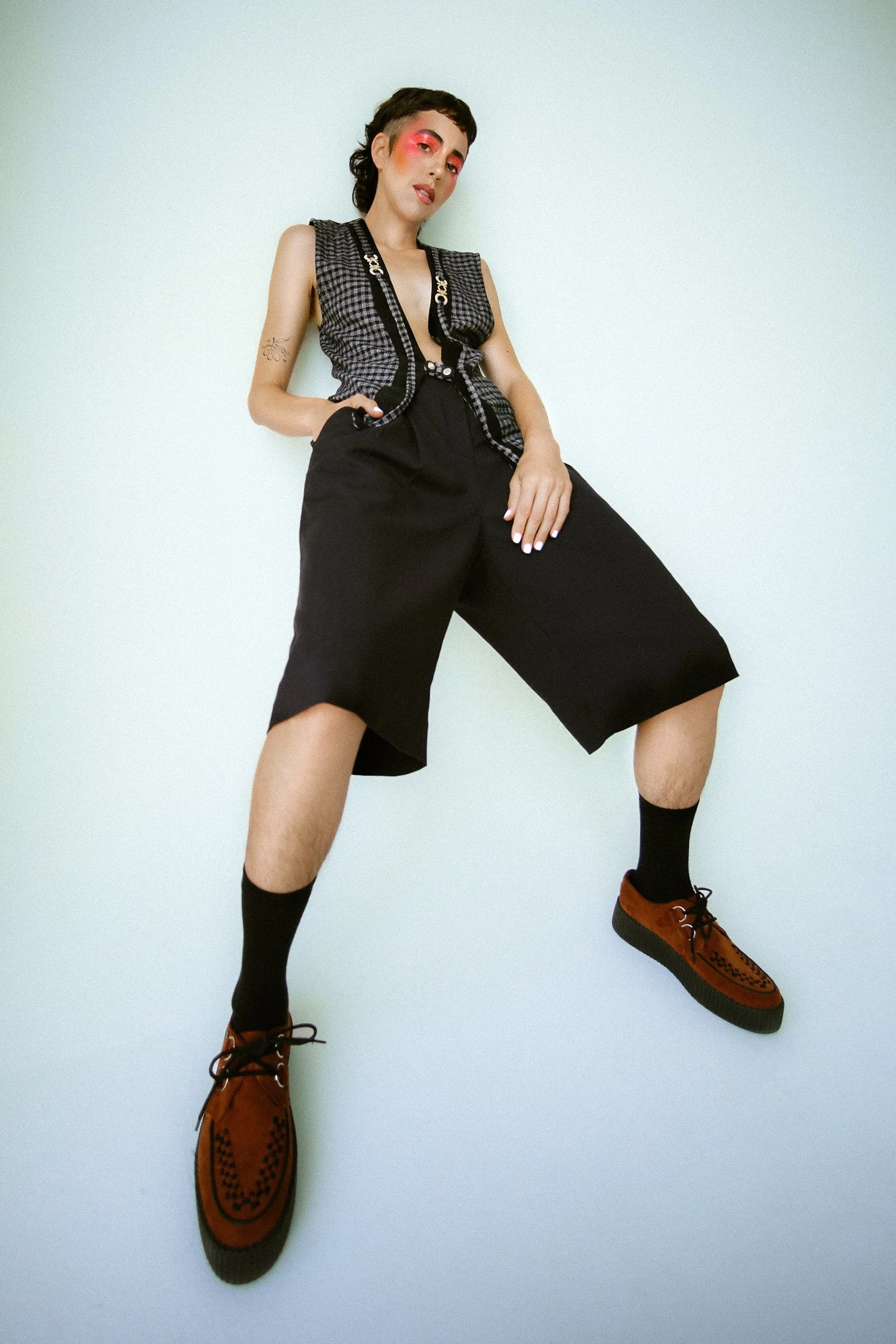 A white Person/model with short hair and colorful makeup, wearing a checkered vest, black shorts, black socks, and brown shoes, posing against a plain white background, shot for an editorial fashion photography session in Los Angeles with fashion pho