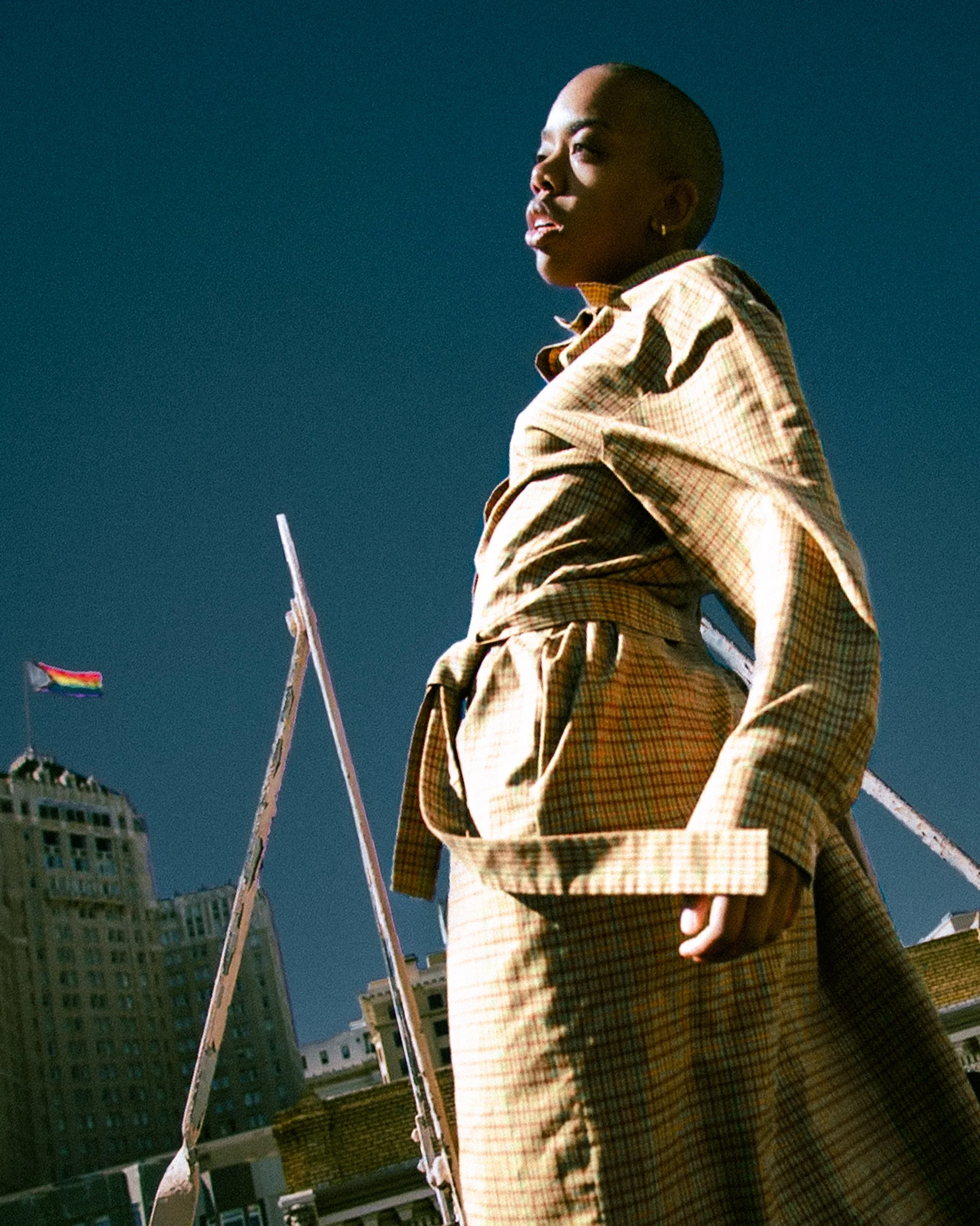 Person with short hair wearing a tan checkered outfit standing outdoors under a clear blue sky, with buildings and a rainbow pride flag in the background, photographed during a photography session in California for a portrait editorial with fashion p