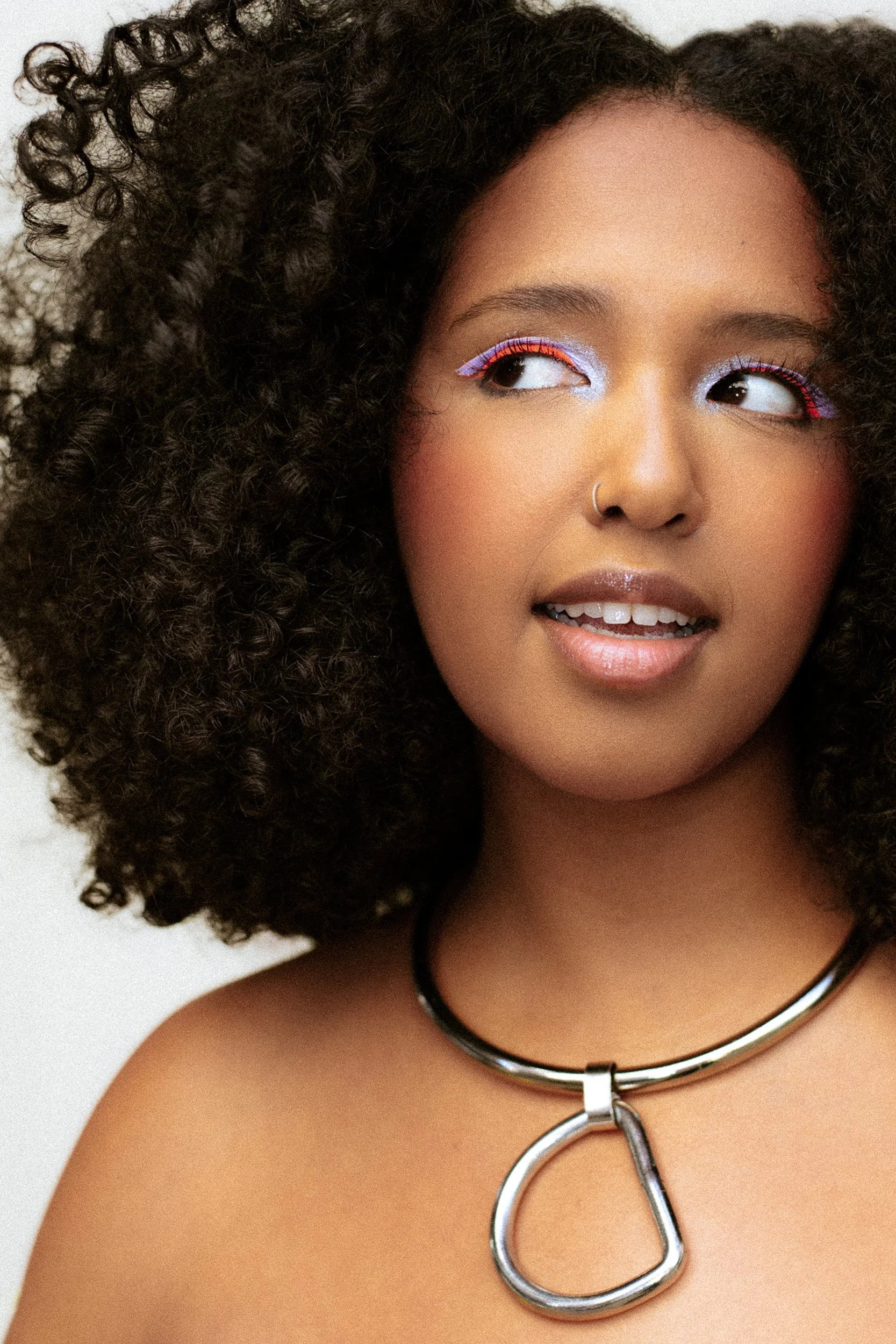 A woman with curly dark hair, colorful eye makeup, a nose ring, and a metallic necklace with a large ring pendant, photographed during a studio photography session in Los Angeles for a fashion editorial collaboration with fashion photographer