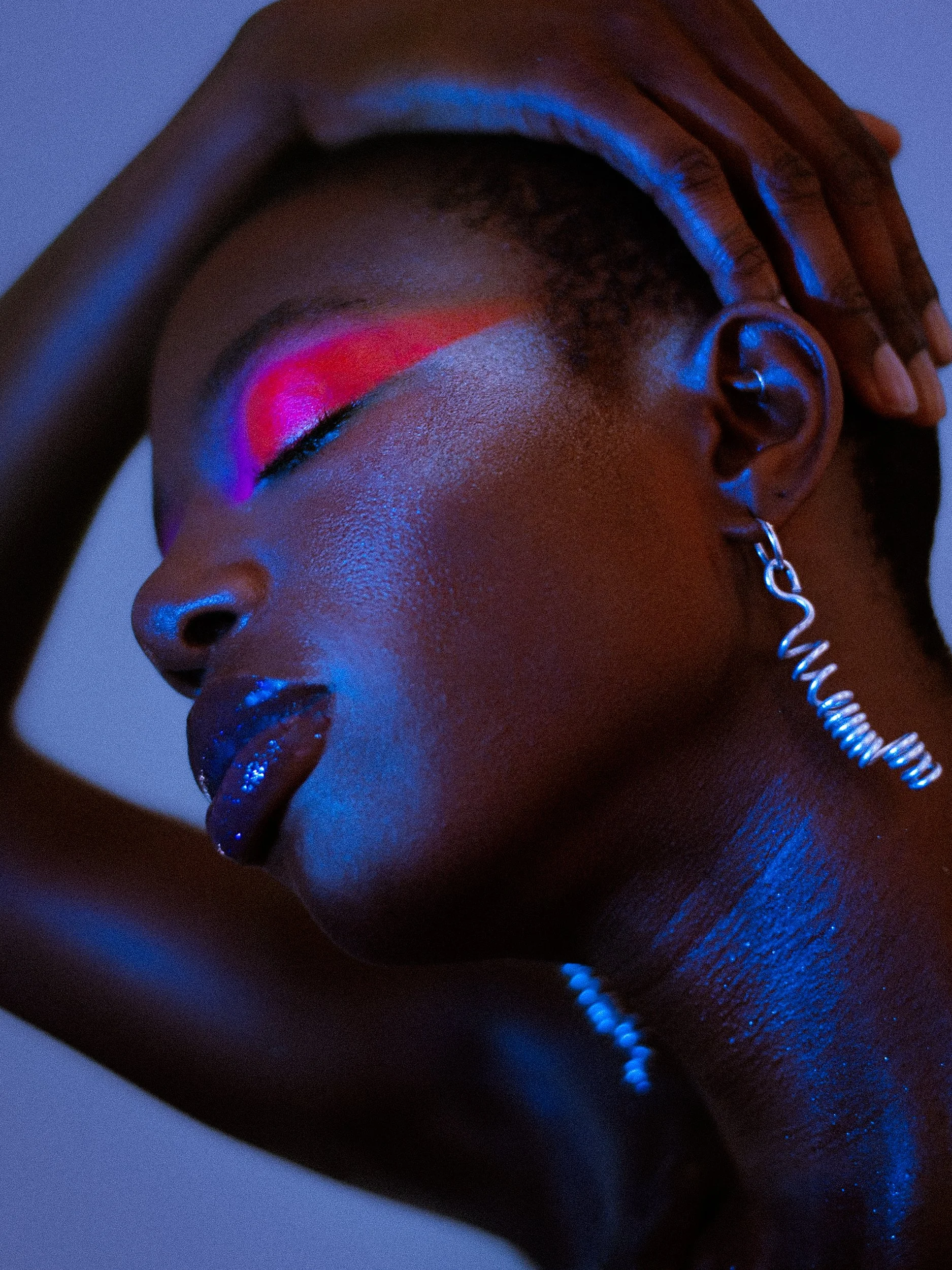A close-up of a woman with vibrant neon makeup, dark lipstick, and a glowing earring, resting her head on a surface with one hand on her forehead, in low blue lighting, A woman with curly black hair wearing bright makeup, earrings, a rainbow beaded b