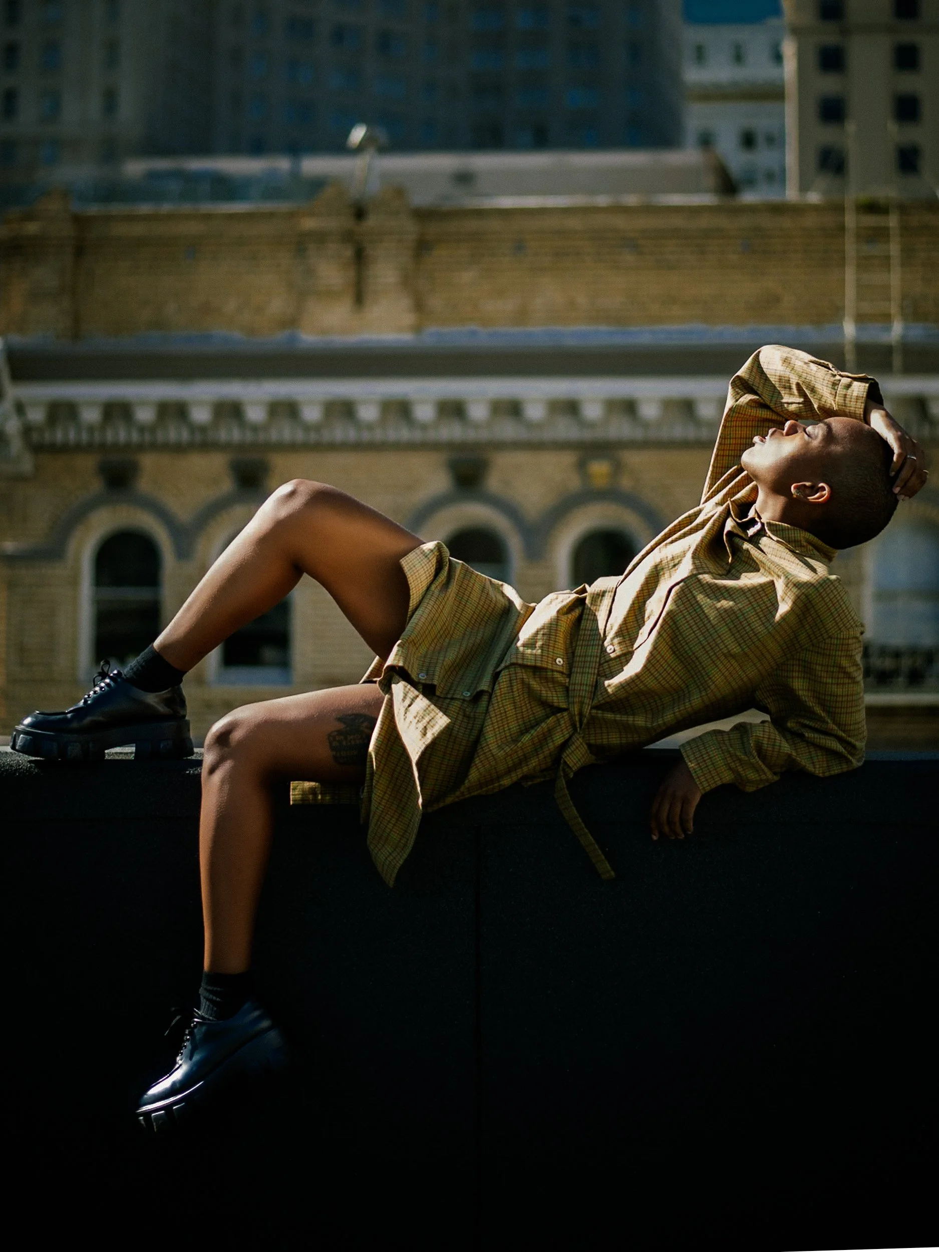 A person wearing a yellow plaid shirt and shorts, lying on a ledge with one hand behind their head, facing upward with eyes closed, against a cityscape background, photographed during a photography session in California for a portrait editorial with 