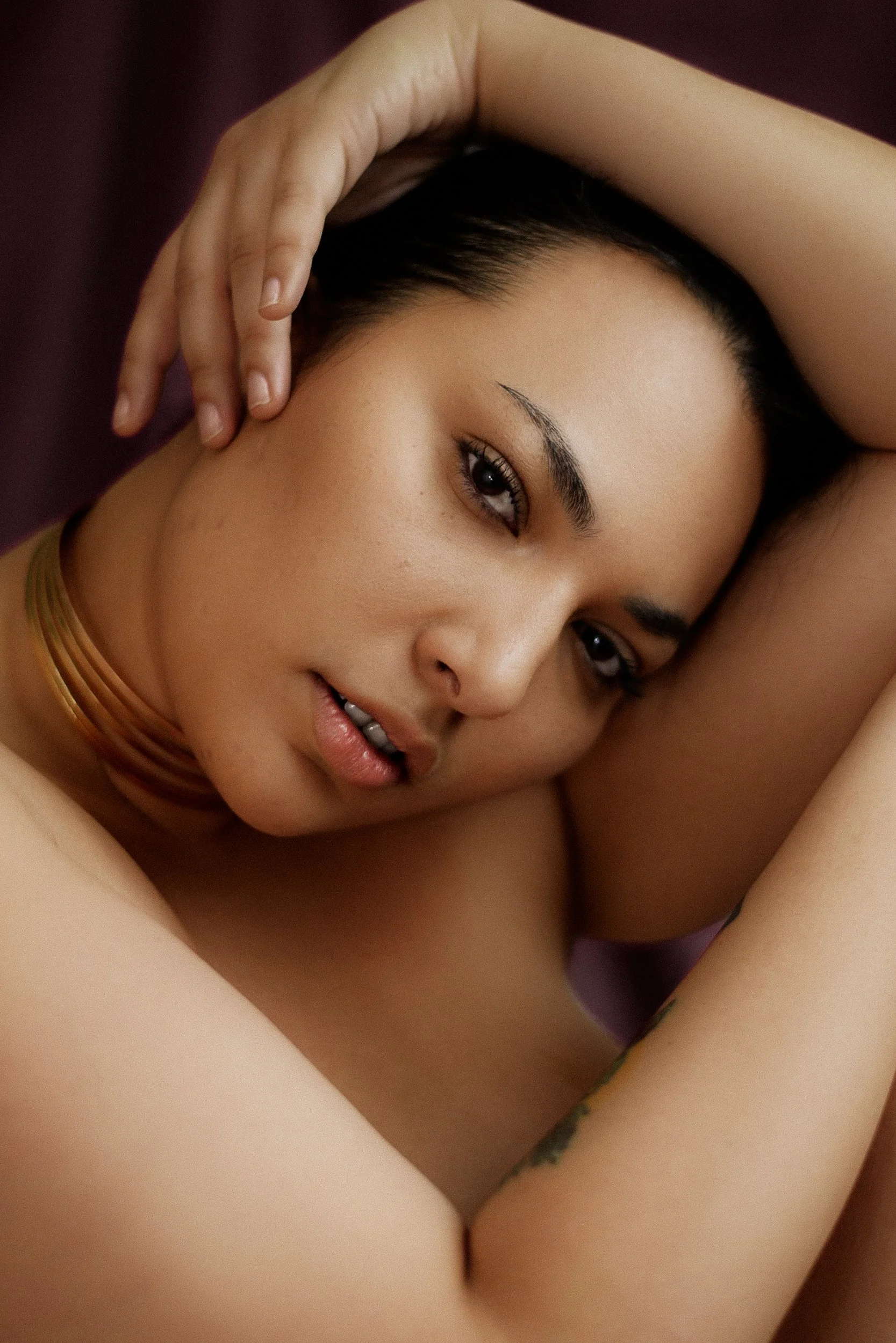A close-up of a woman with dark hair, lying down with one arm raised over her head, gazing into the camera with a relaxed expression, wearing a gold choker necklace, photographed during a jewelry editorial photography session in Los Angeles with fash