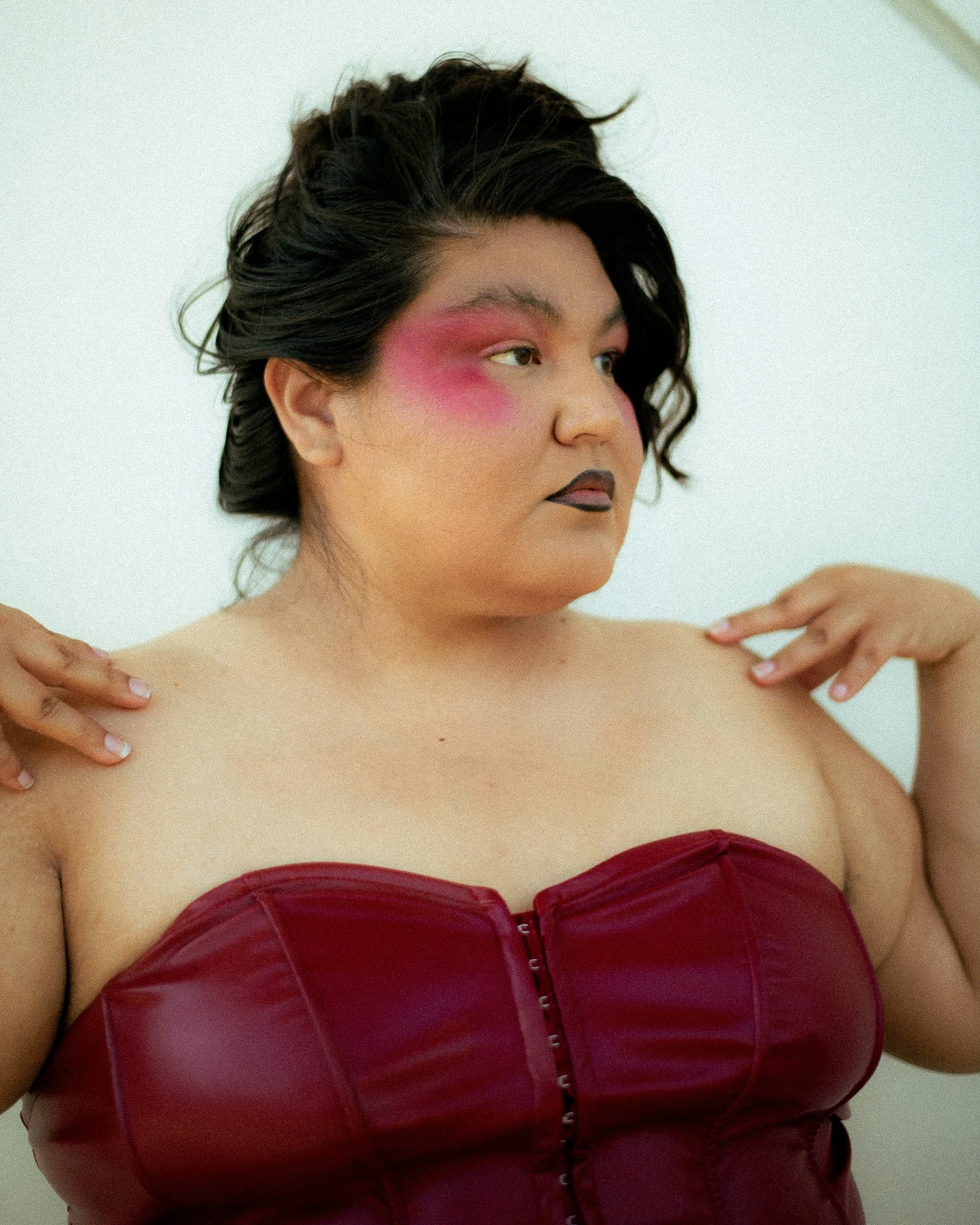 A Latina woman model with bold makeup, wearing a red strapless top, touching shoulders against a plain background, photographed during a fashion editorial photography session in Los Angeles with fashion photographer Gí Tenamá...